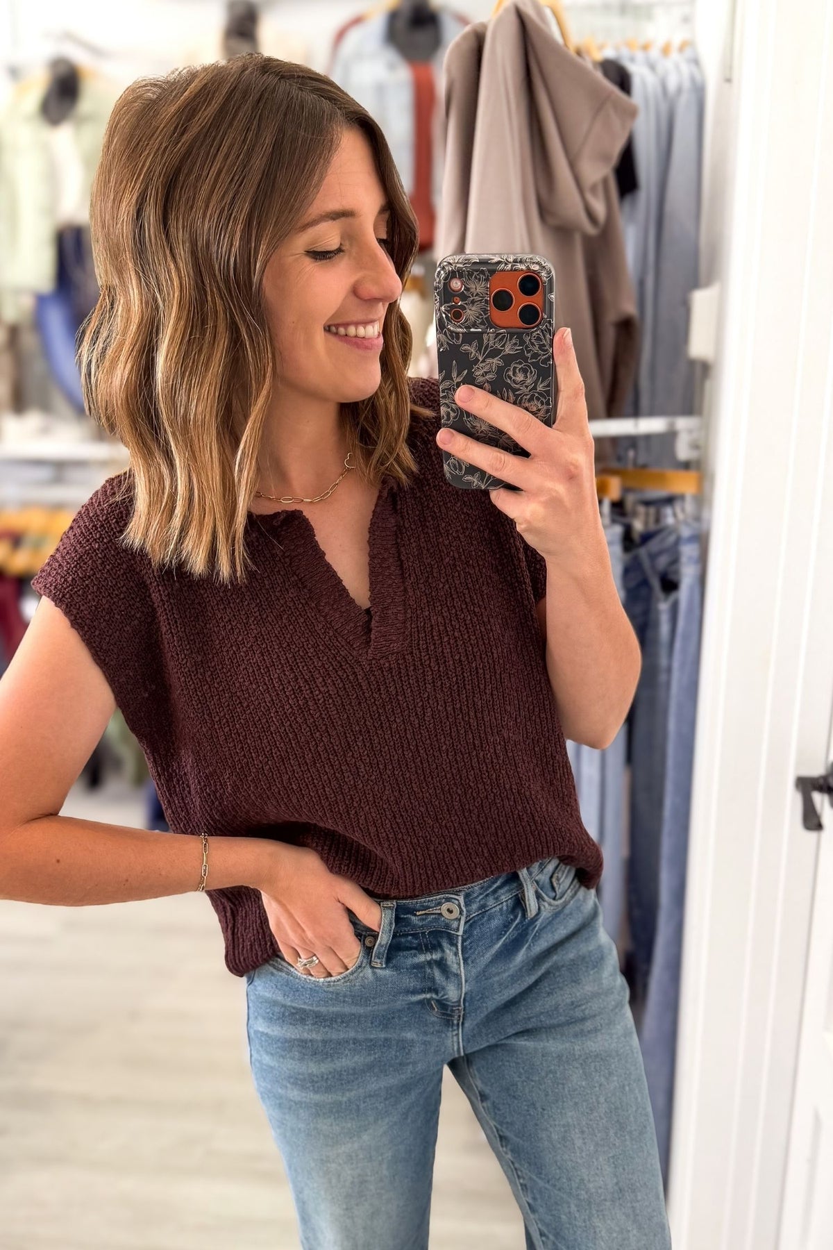 A woman takes a mirror selfie in a clothing store, wearing a short-sleeve chocolate knit top and blue jeans.