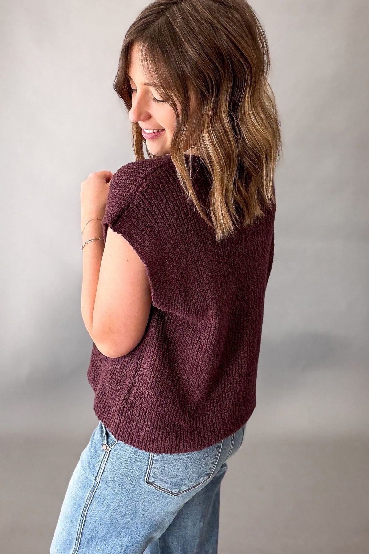 A woman wearing a short-sleeve, textured knit top in chocolate, paired with light blue jeans, photographed against a neutral gray studio background.