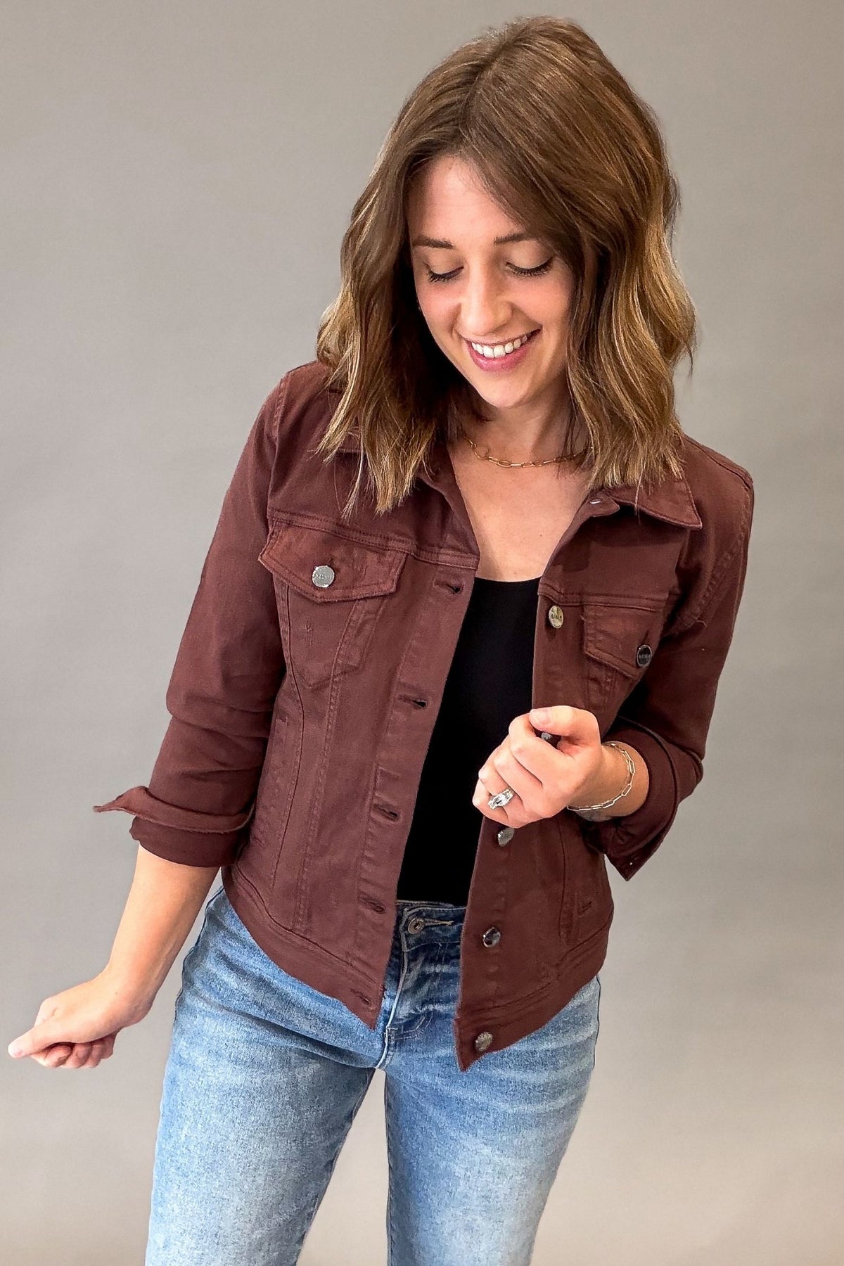A woman wearing a chocolate utility jacket over a black top and blue jeans, standing against a plain gray studio background.