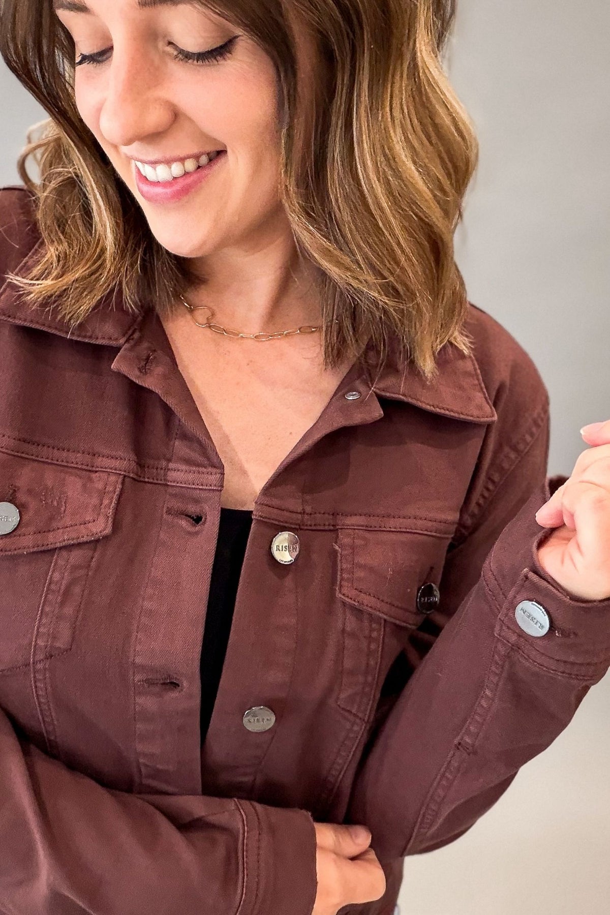 A woman wearing a rich chocolate utility jacket with silver buttons, layered over a black top, shown in a close-up shot against a neutral gray background.