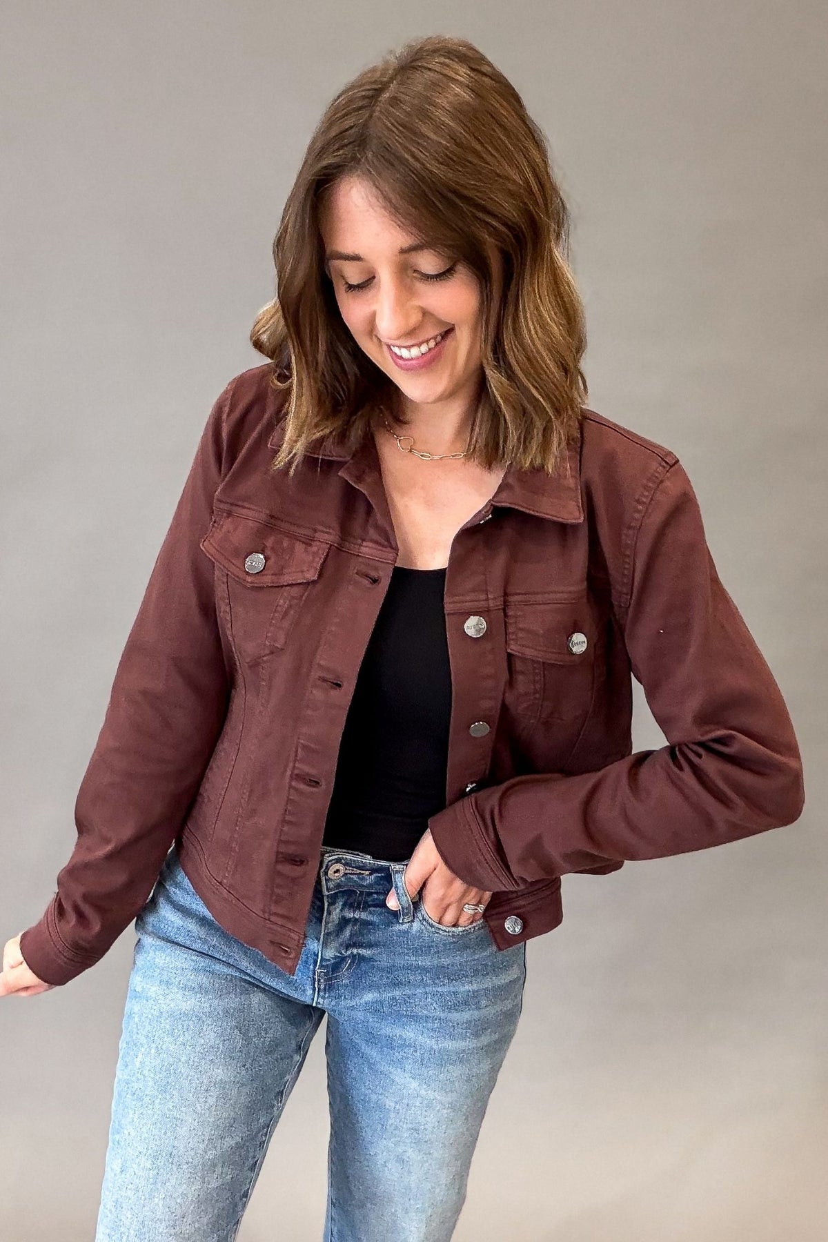 A woman wearing a long-sleeve rich chocolate utility jacket over a black top and light-wash blue jeans, posing against a neutral gray studio background.