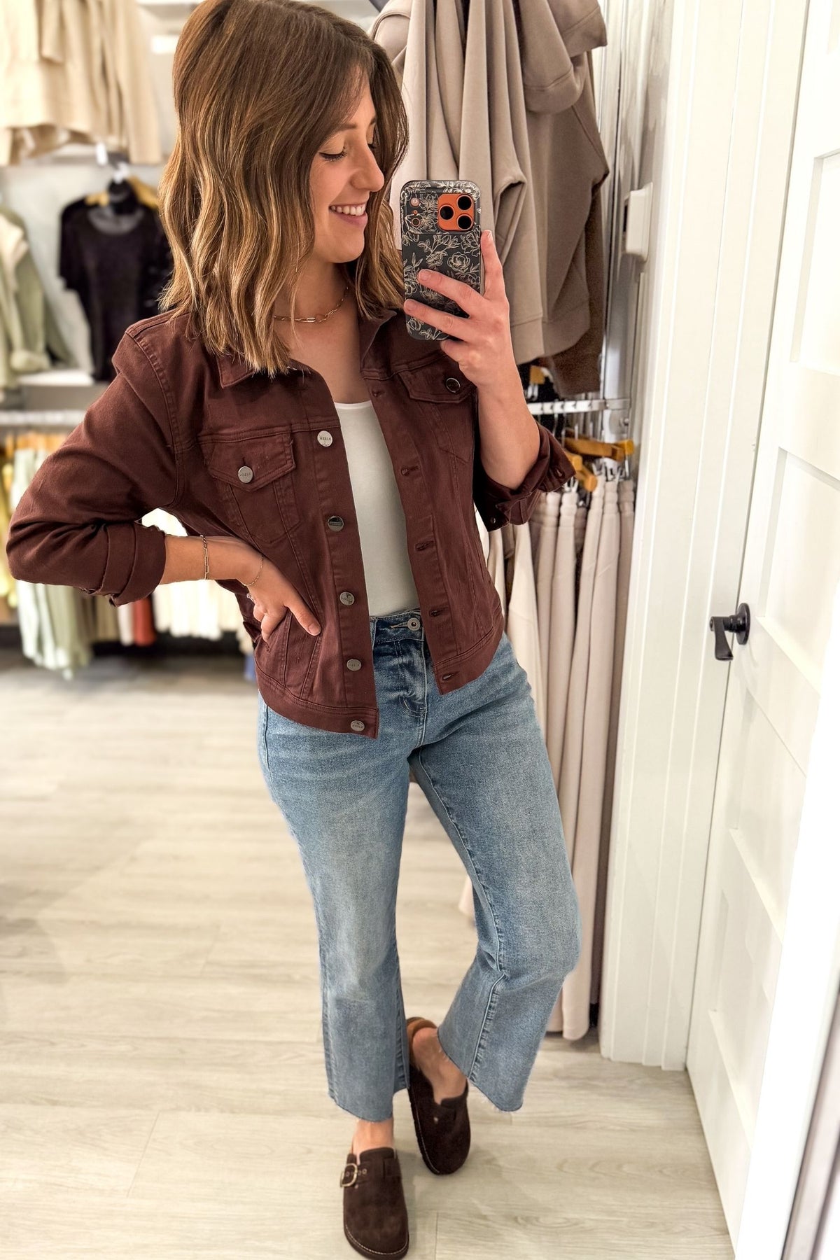 A woman takes a mirror selfie in a clothing store, wearing a chocolate utility button-up jacket over a white top, light-wash blue jeans, and brown loafers.