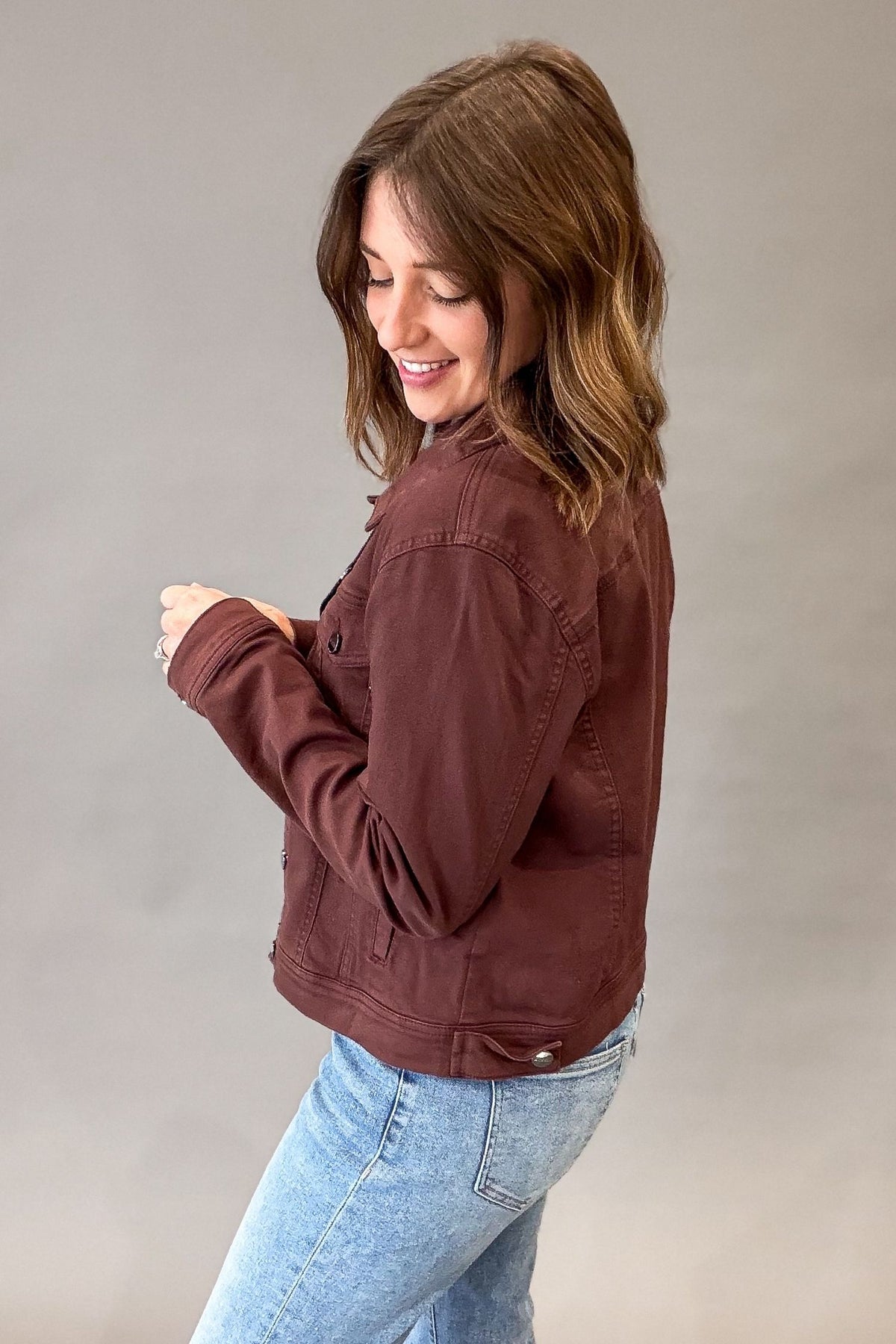 A woman wearing a rich chocolate utility jacket and light blue jeans, photographed against a plain gray studio background.