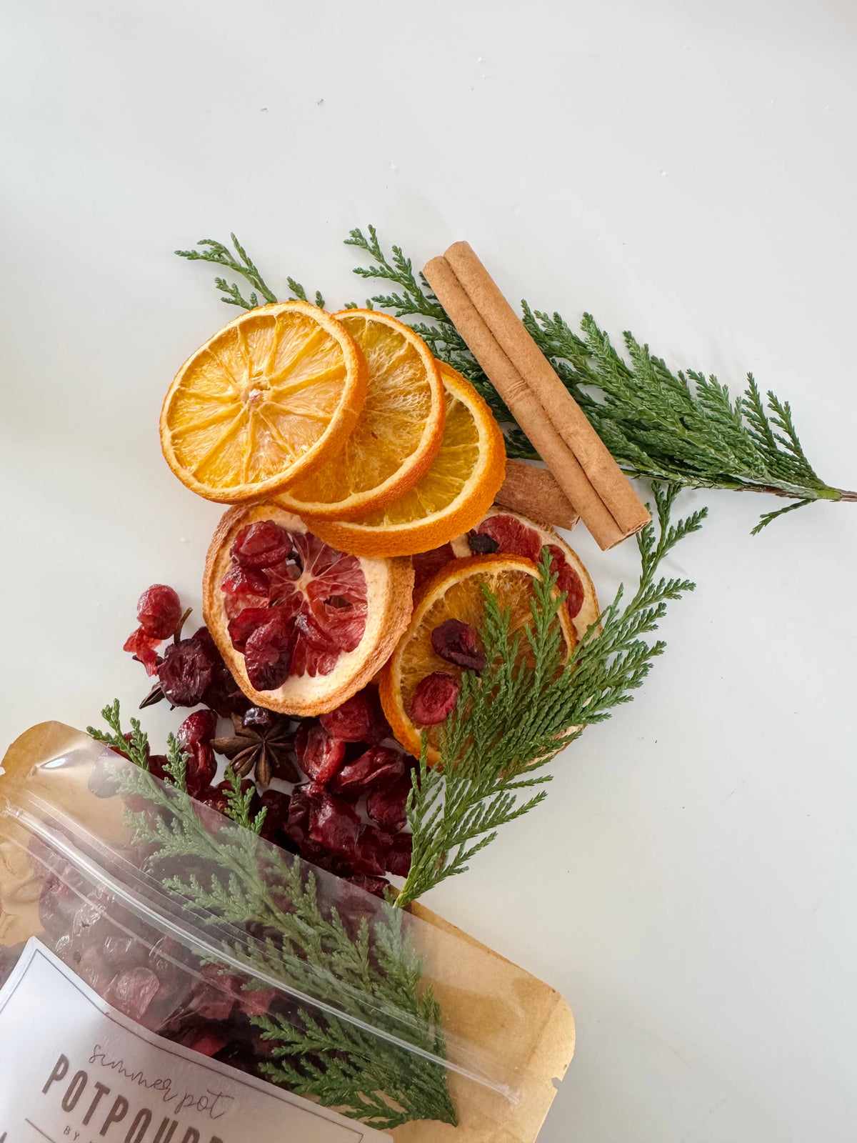 Dried oranges, cinnamon sticks, and pine needles on a white surface with a labeled potpourri bag.