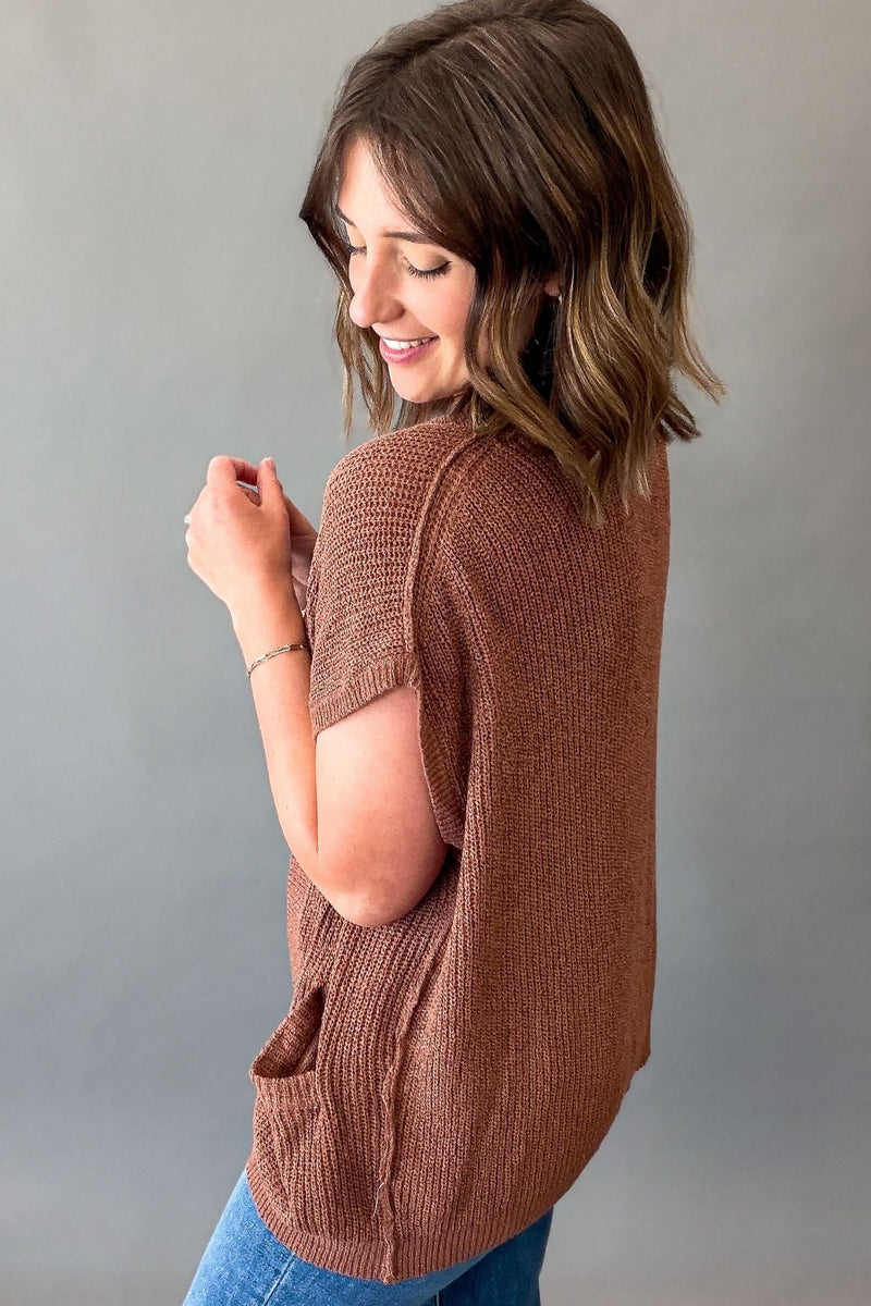 A woman wearing a brown short-sleeve knit top with side pockets, shown from the back against a plain gray background.