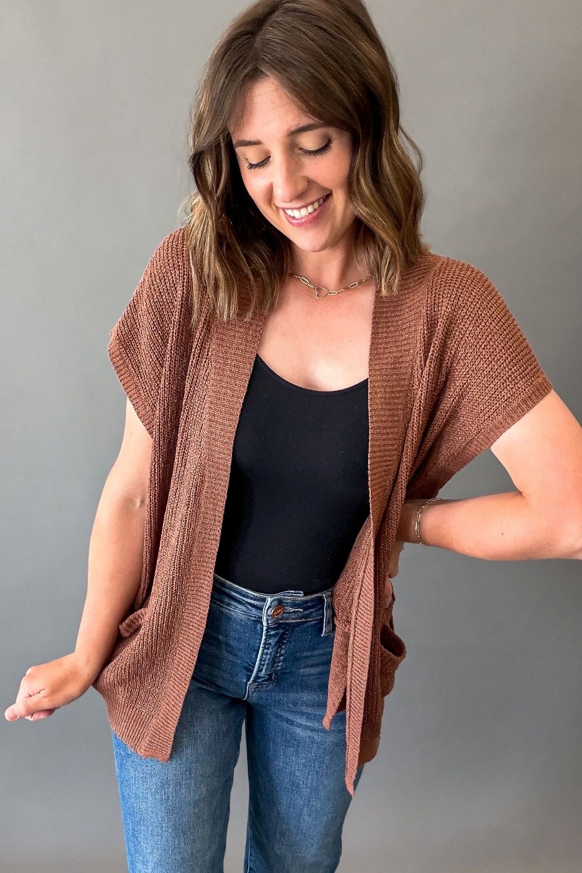 A woman wearing a short-sleeve, open-front knit cardigan in a warm brown color, styled over a black tank top and blue jeans, posing against a solid gray background.