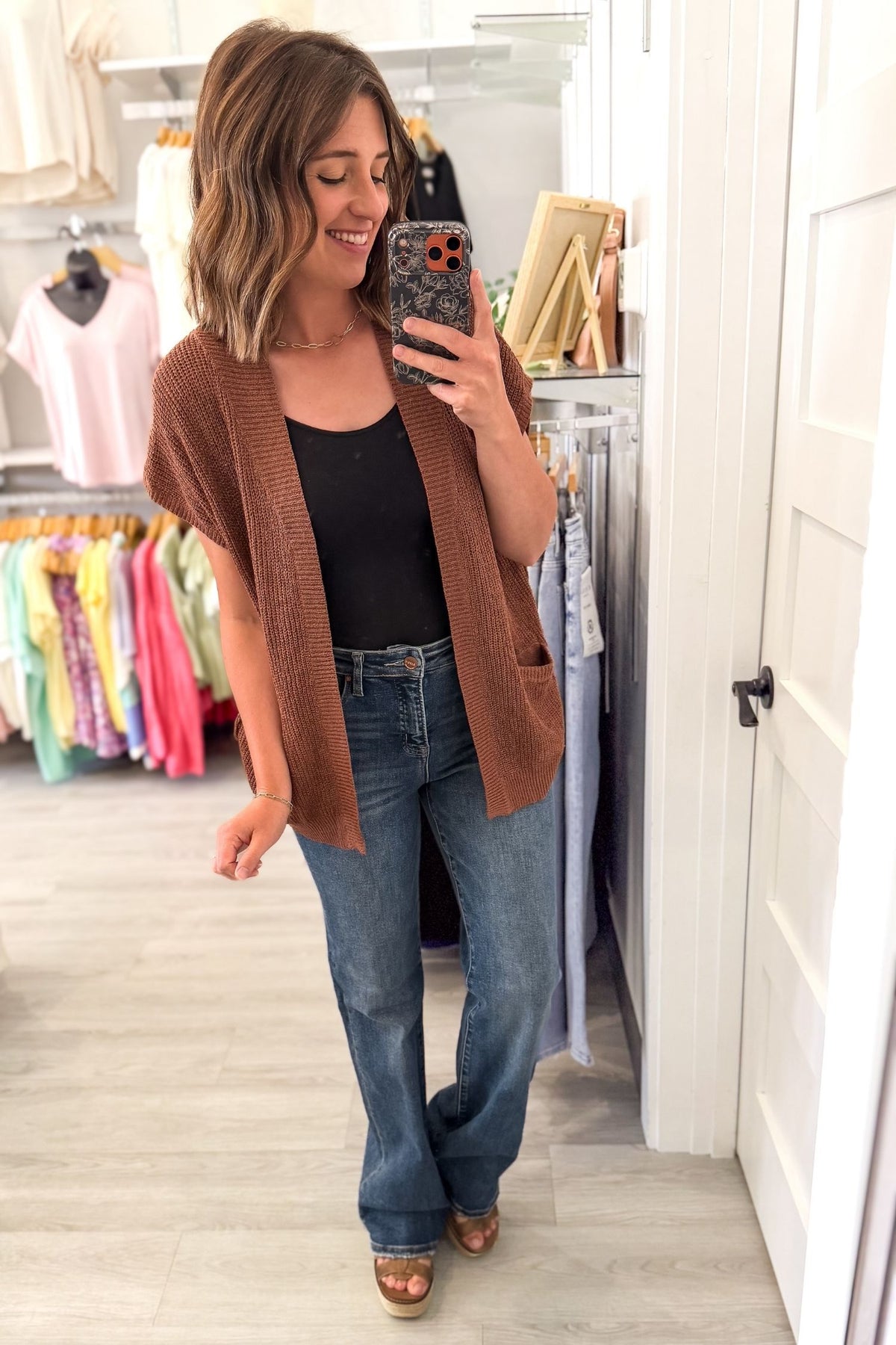 A woman takes a mirror selfie in a clothing boutique, wearing a brown open-front knit cardigan over a black top and blue jeans.