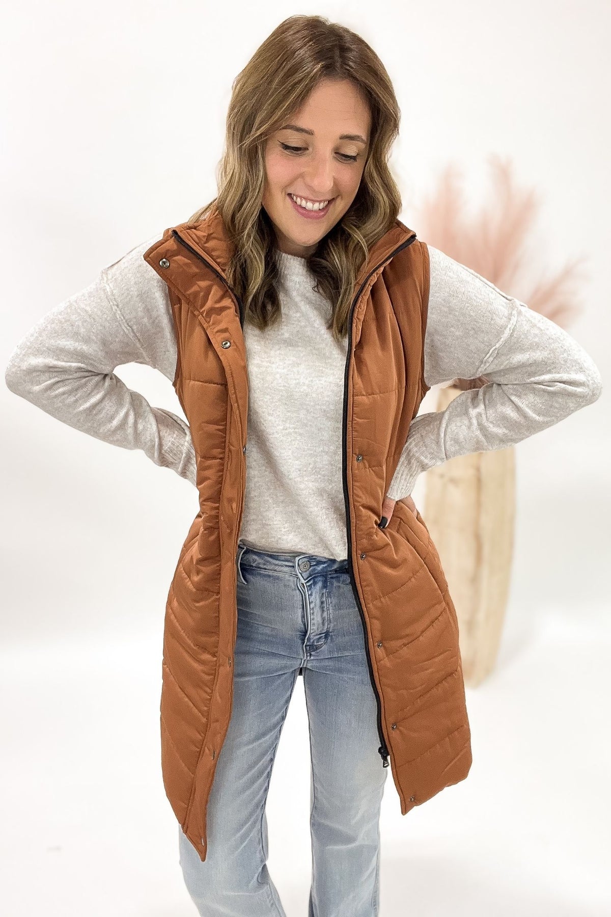 Woman wearing a brown puffer vest over a light gray sweater and blue jeans on a white background