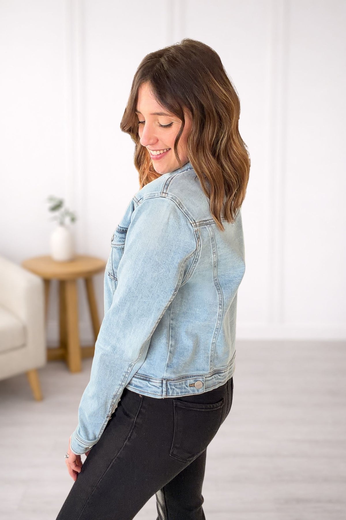 Woman wearing a light blue denim jacket in a room with a white wall and a small table.