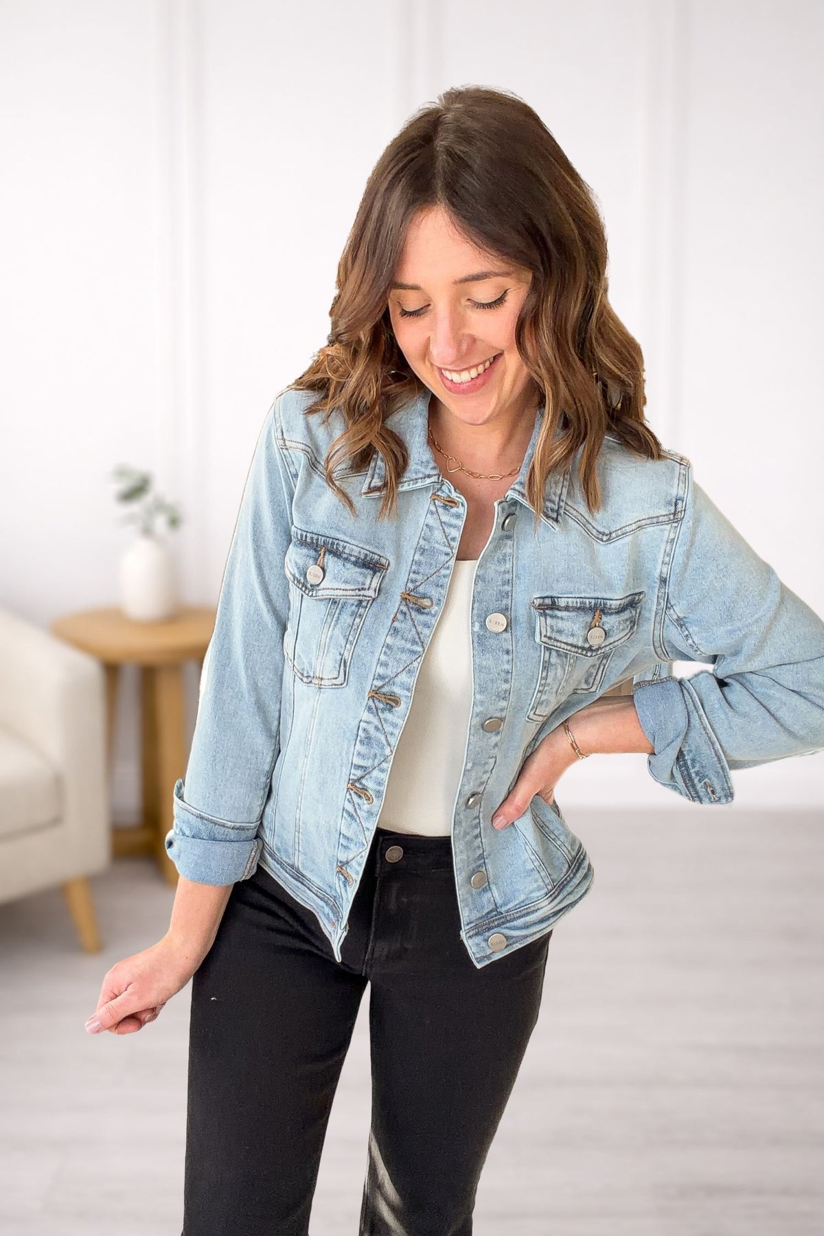Woman wearing a light blue denim jacket with black jeans in a room with a white wall and a small table.
