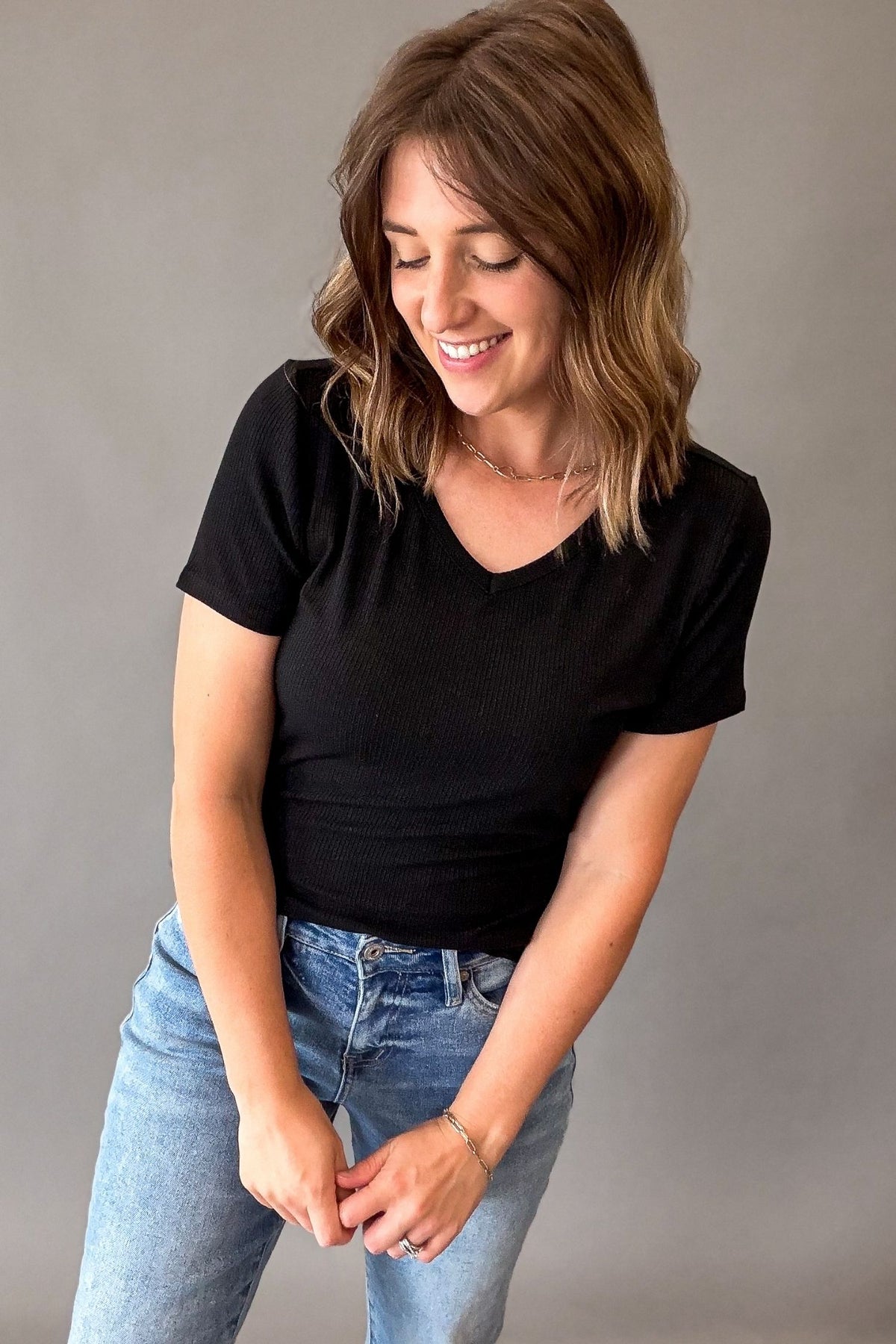 A woman with shoulder-length brown hair is wearing a fitted black short-sleeve V-neck ribbed t-shirt and light-wash blue jeans, standing against a neutral gray studio background.
