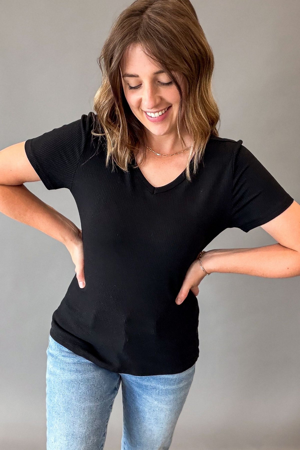 A woman wearing a black short-sleeve V-neck ribbed knit top and light blue jeans, posing with her hands on her hips against a plain gray studio background.