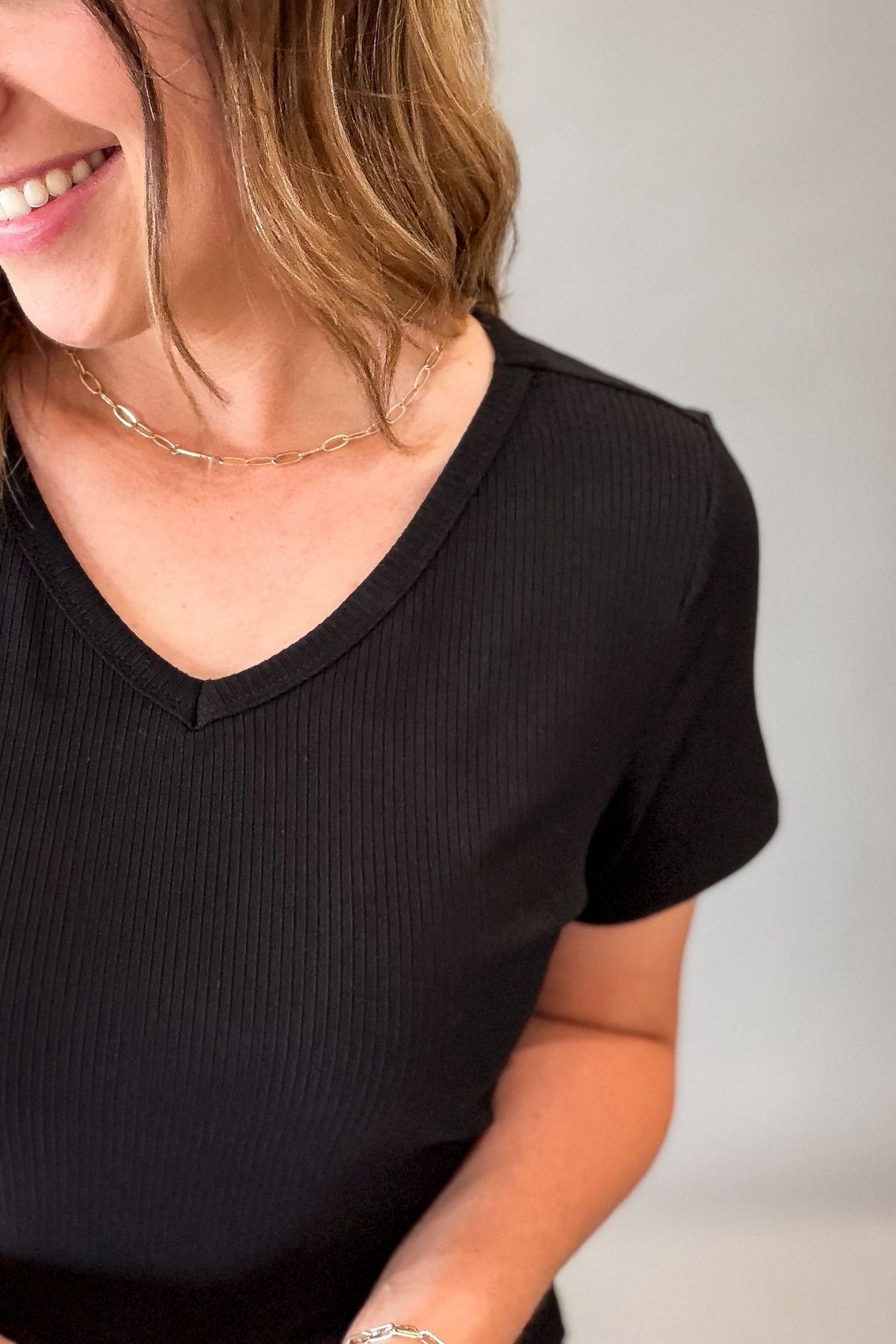 A close-up of a woman wearing a black ribbed short-sleeve V-neck top, showing her smiling and neck with a delicate gold chain necklace.
