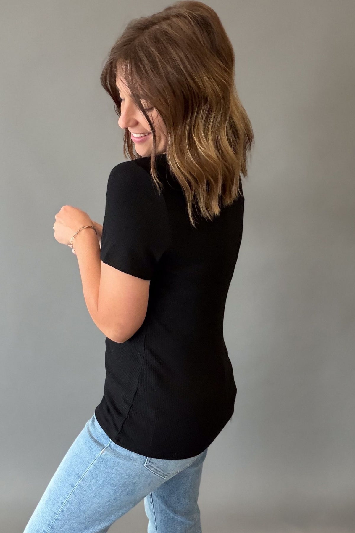 A woman with shoulder-length brown hair is shown from the back wearing a fitted black short-sleeve top and light blue jeans against a plain gray studio background.