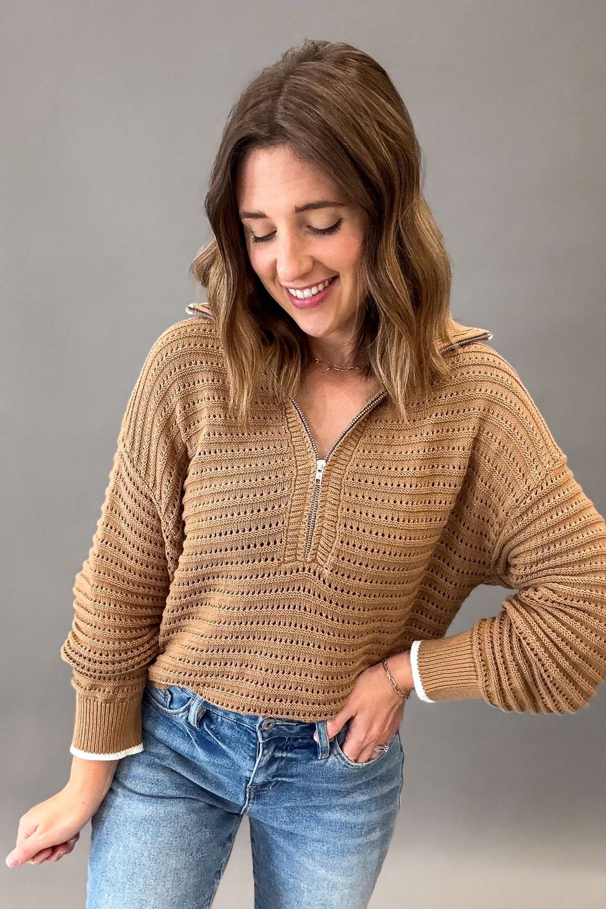 A woman wearing a coconut colored ribbed knit half-zip sweater with a half-placket and ribbed cuffs, paired with blue jeans, posing against a solid gray studio background.