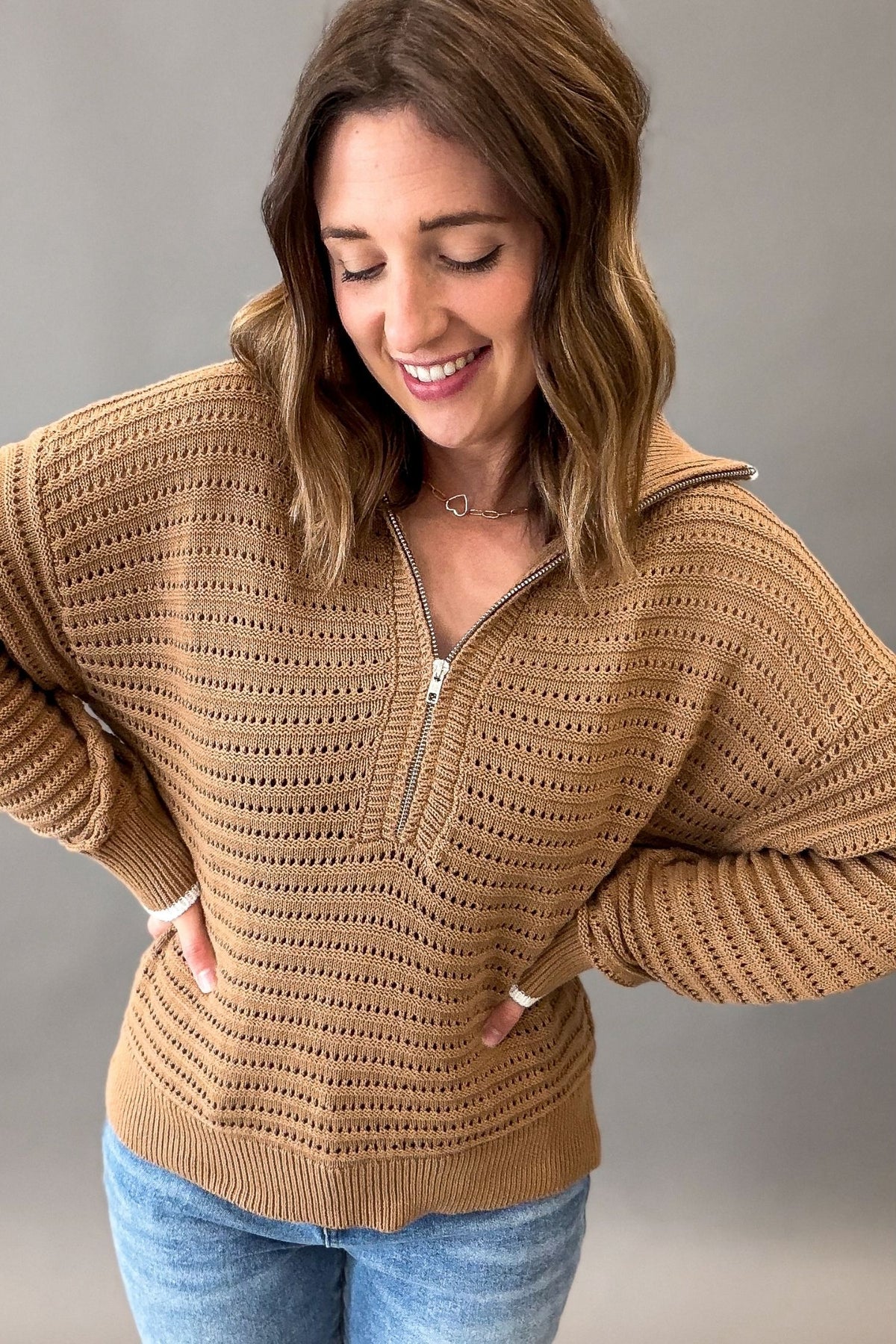 A woman wearing a coconut colored ribbed knit half-zip sweater with a high collar and front pockets, paired with blue jeans, photographed against a neutral gray studio background.