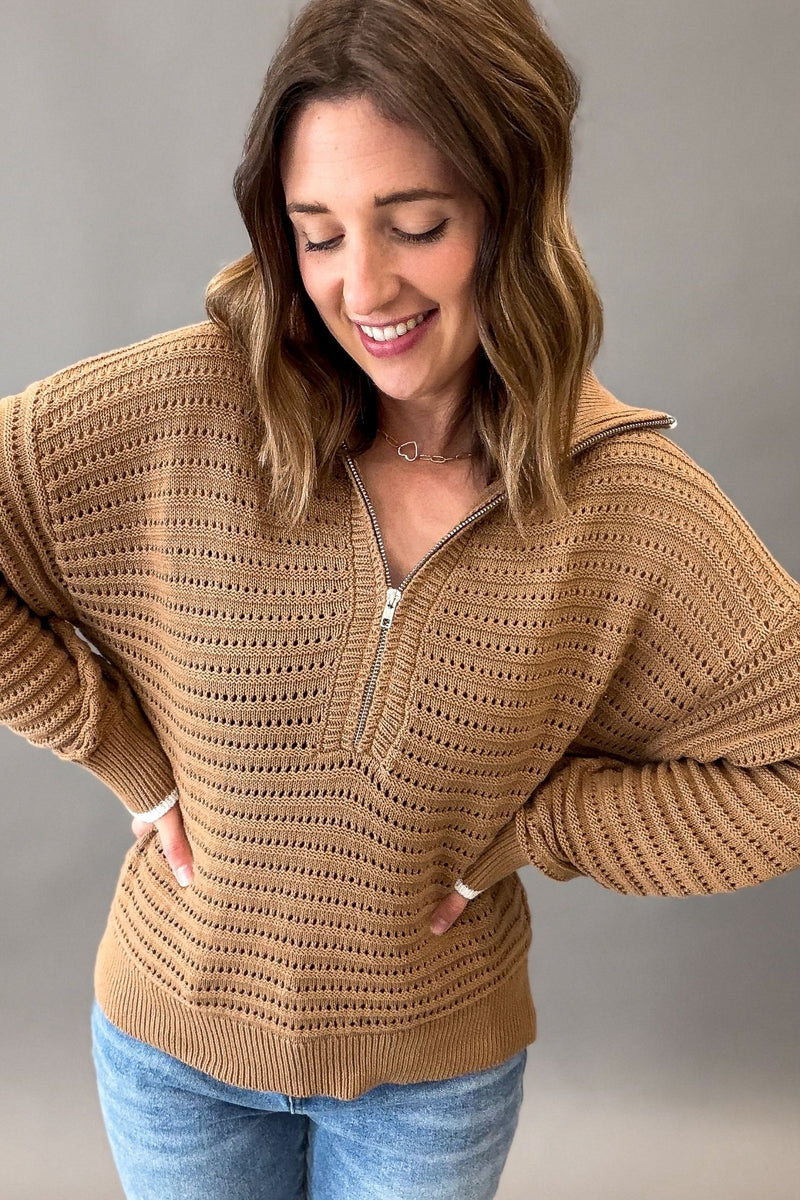 A woman wearing a coconut colored ribbed knit half-zip sweater with a high collar and front pockets, paired with blue jeans, photographed against a neutral gray studio background.