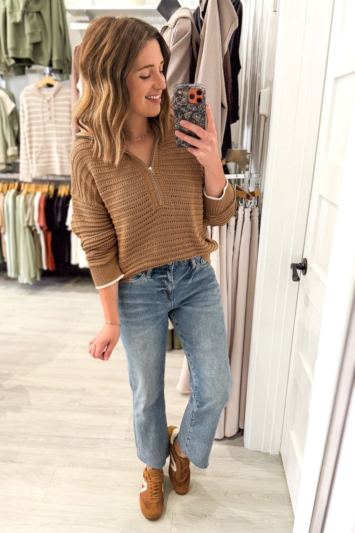 A woman takes a mirror selfie in a clothing store wearing a coconut textured quarter-zip knit sweater, light-wash blue jeans, and brown suede boots.