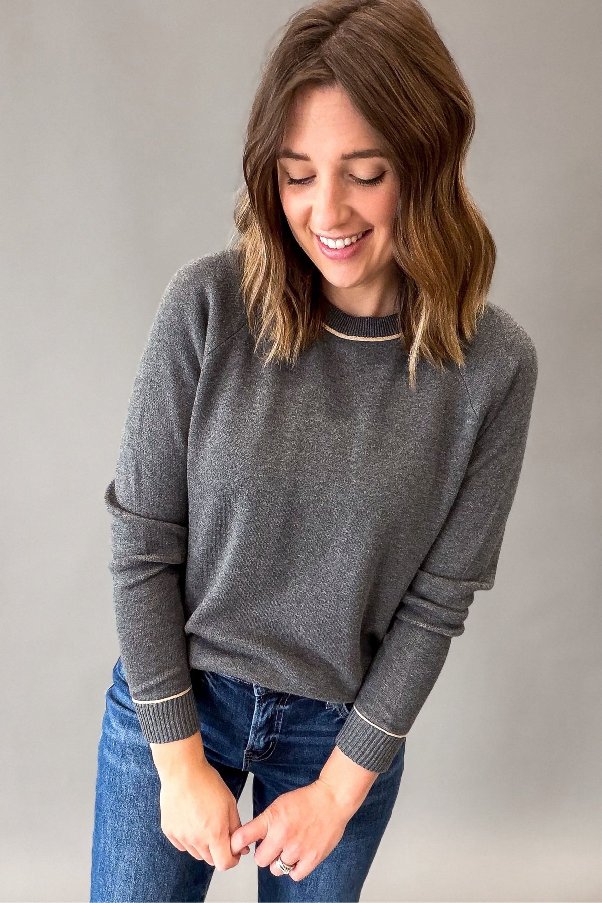 A woman wearing a dark gray long-sleeve knit sweater with ribbed cuffs and a thin taupe piping along neckline and cuffs, paired with blue jeans, standing against a neutral gray background.