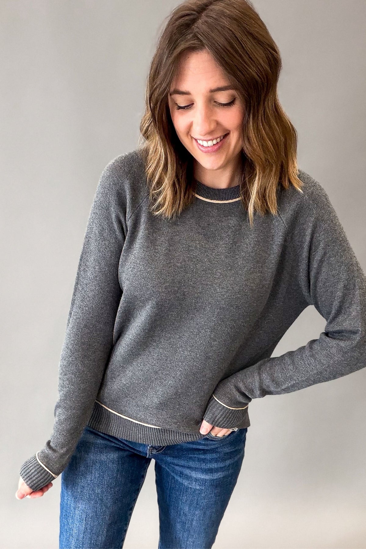 A woman wearing a charcoal crewneck sweater with thin cream-colored trim at the collar, cuffs, and hem, paired with blue jeans, photographed against a neutral gray background.
