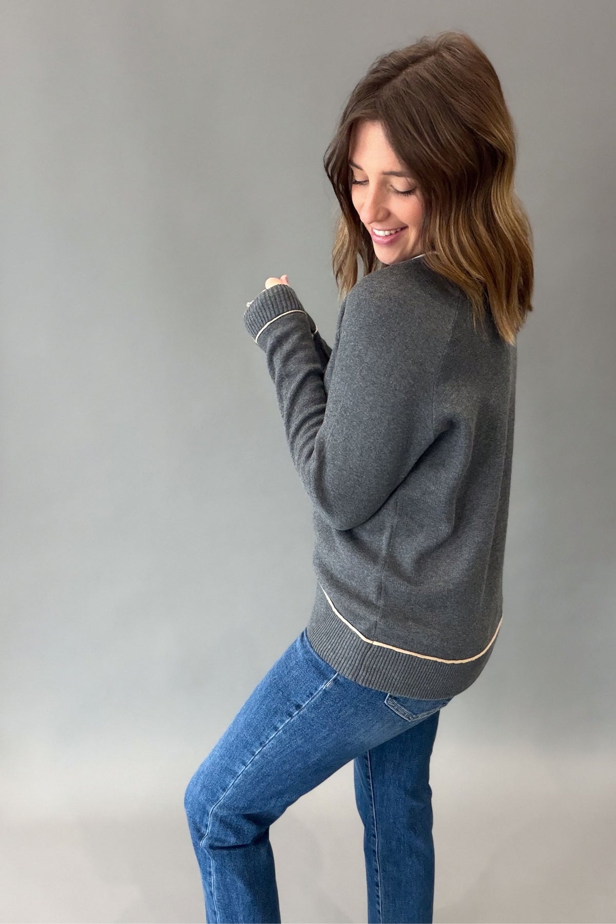 A woman wearing a dark gray knit sweater with cream ribbed trim and blue jeans, posing against a neutral gray studio background.