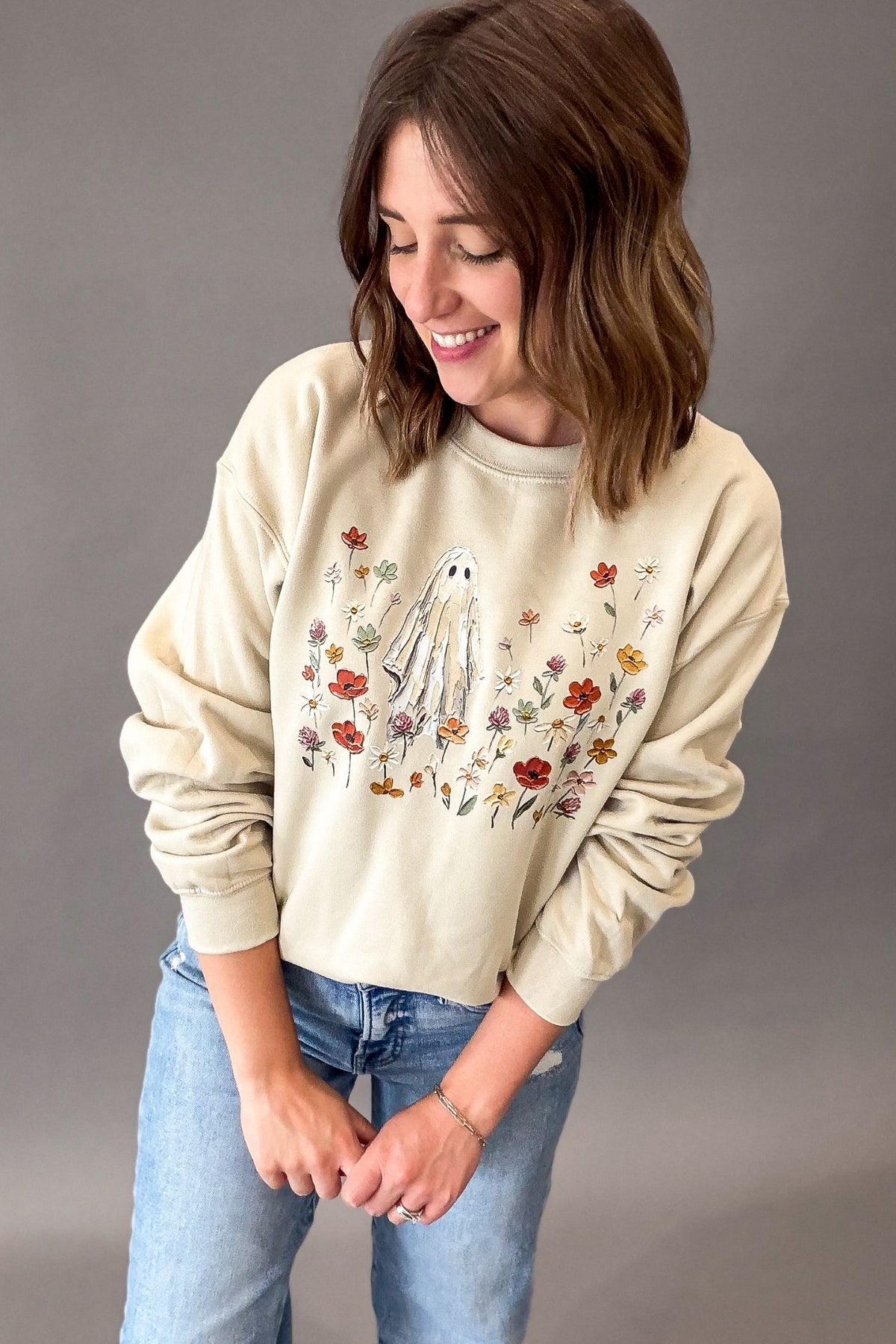 A woman wearing a beige-colored crewneck sweatshirt with a ghost and floral graphic, paired with blue jeans, standing against a solid gray background.