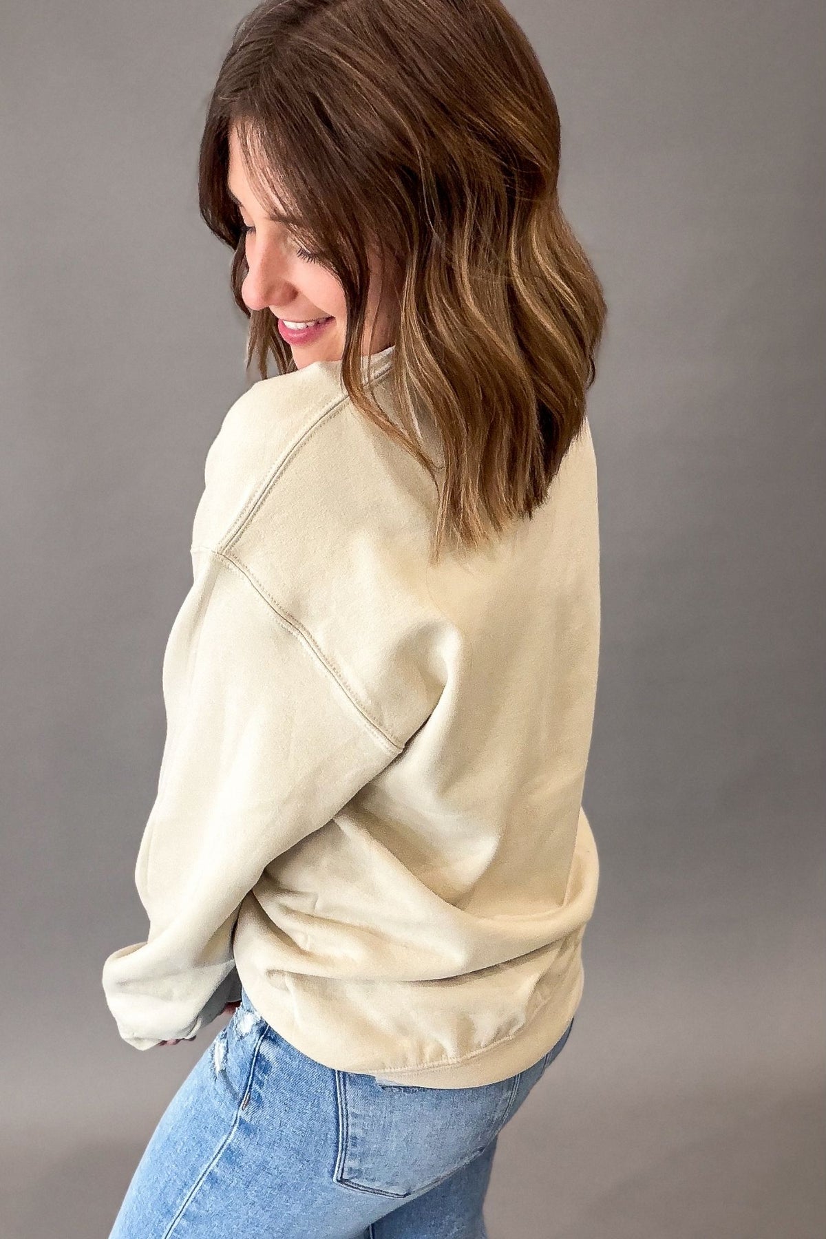A woman with shoulder-length brown hair is shown from the back and side, wearing a beige-colored crewneck sweatshirt and light blue jeans against a solid gray studio background.