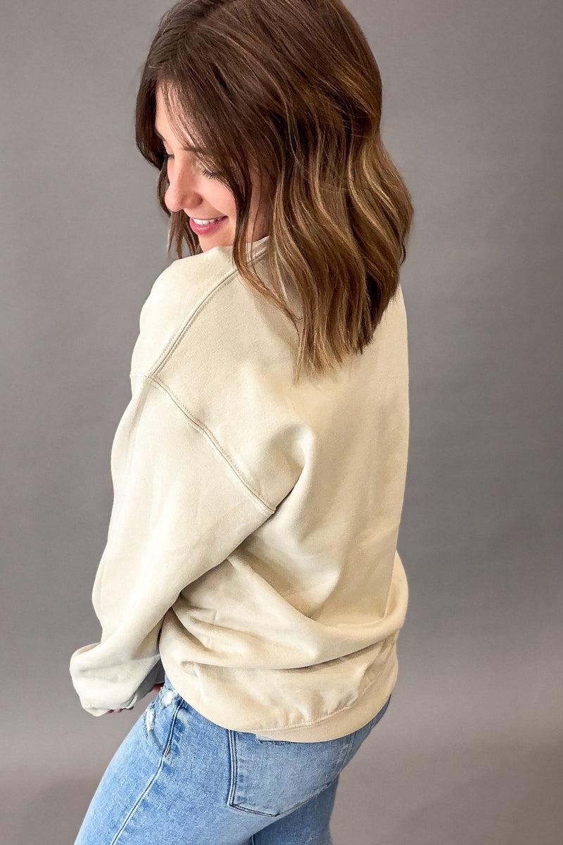 A woman with shoulder-length brown hair is shown from the back and side, wearing a beige-colored crewneck sweatshirt and light blue jeans against a solid gray studio background.