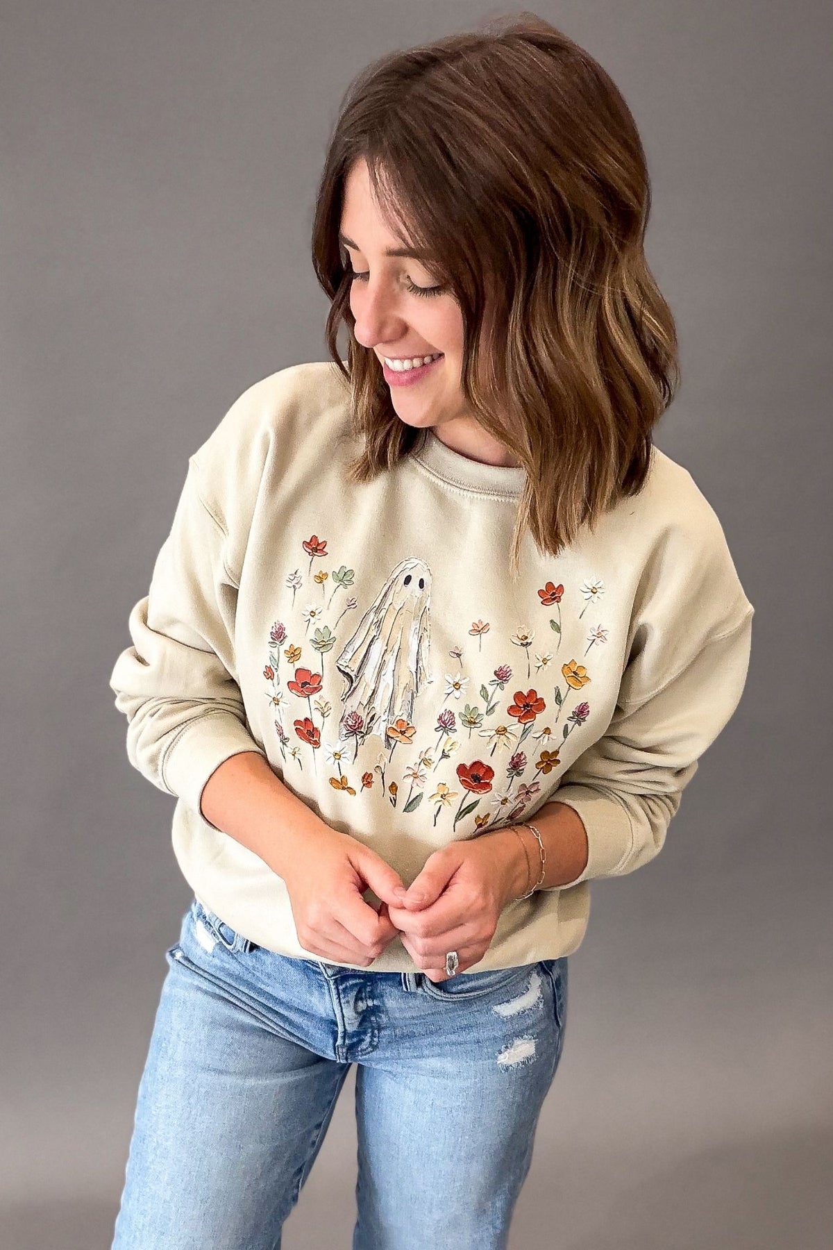 A woman wearing a beige-colored crewneck sweatshirt with a ghost and floral graphic, paired with light-wash blue jeans, standing against a solid gray studio background.