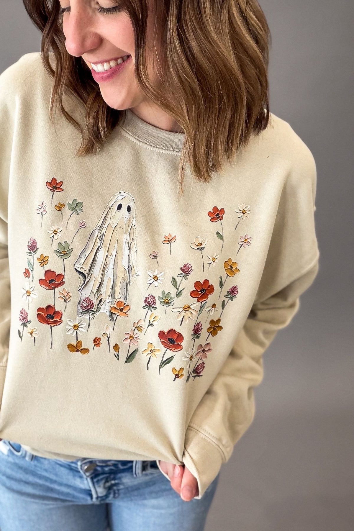 A woman wearing a beige crewneck sweatshirt with a graphic of a white ghost surrounded by colorful wildflowers, paired with blue jeans against a neutral gray background.