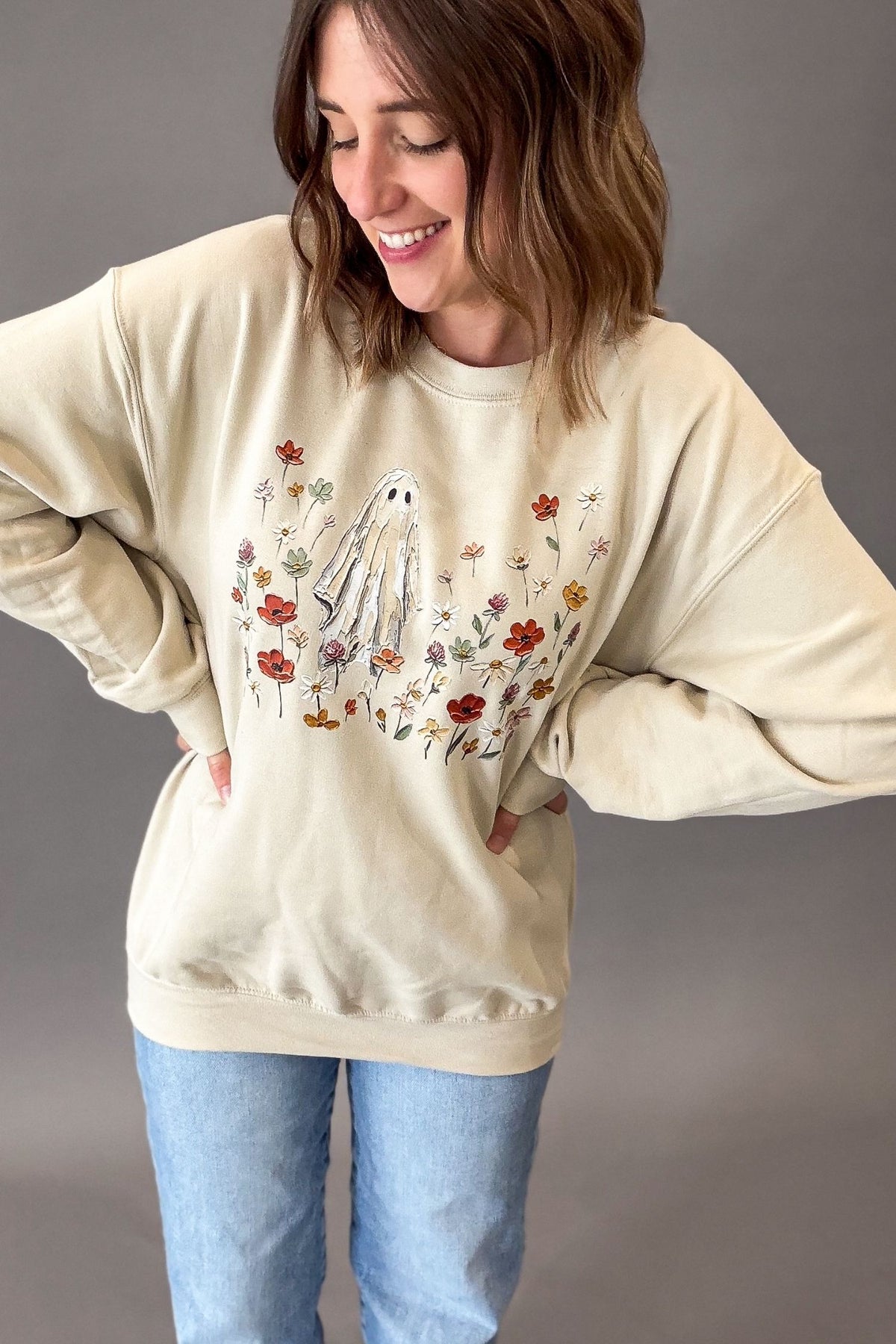 A woman wearing a beige-colored crewneck sweatshirt with a graphic of a white ghost surrounded by red, orange, and purple flowers, paired with light blue jeans, set against a plain gray studio background.