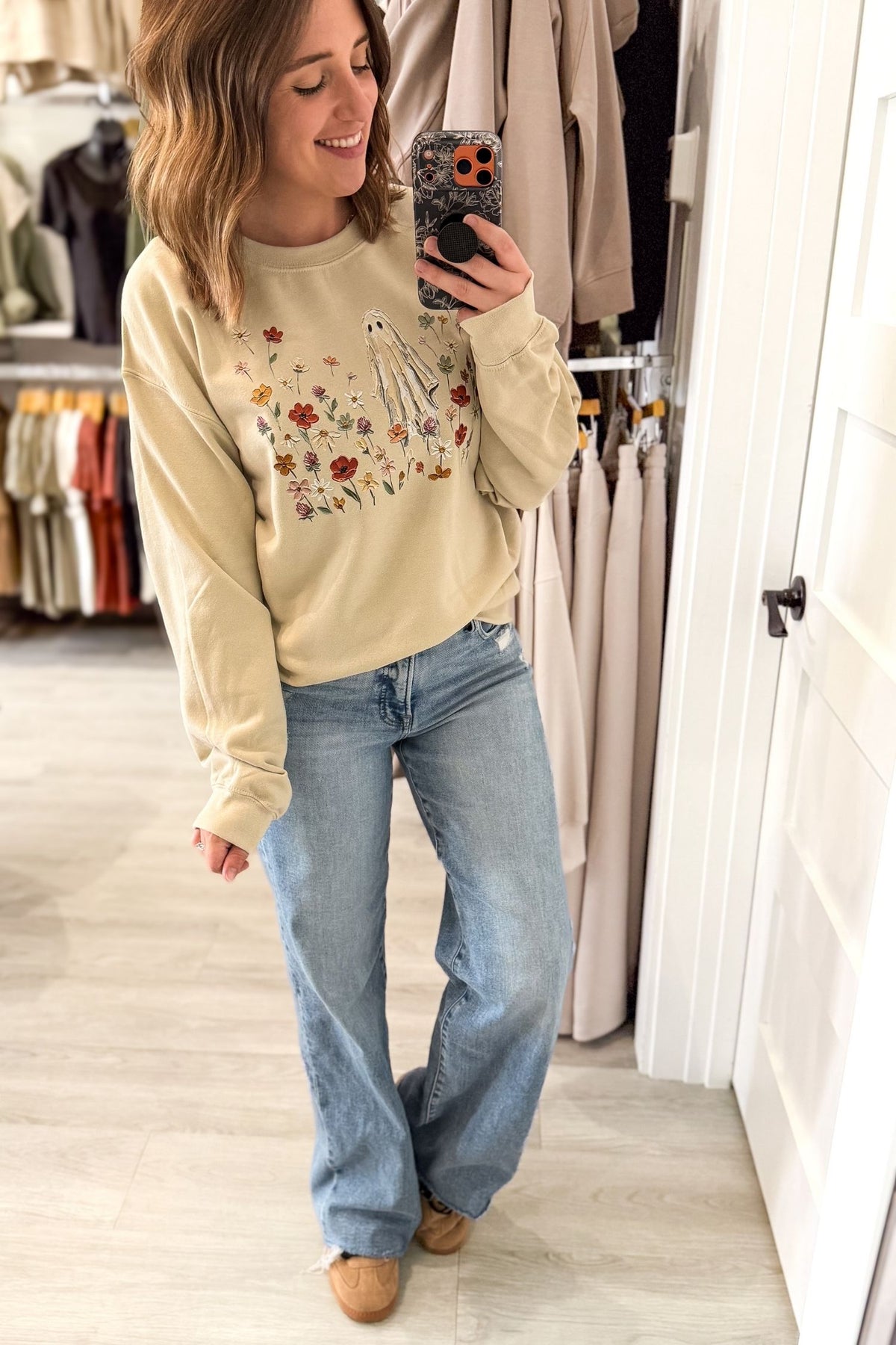 A woman takes a mirror selfie in a clothing boutique wearing a beige crewneck sweatshirt with a white bird and colorful floral graphic paired with light-wash blue jeans and camel corkys sneakers.