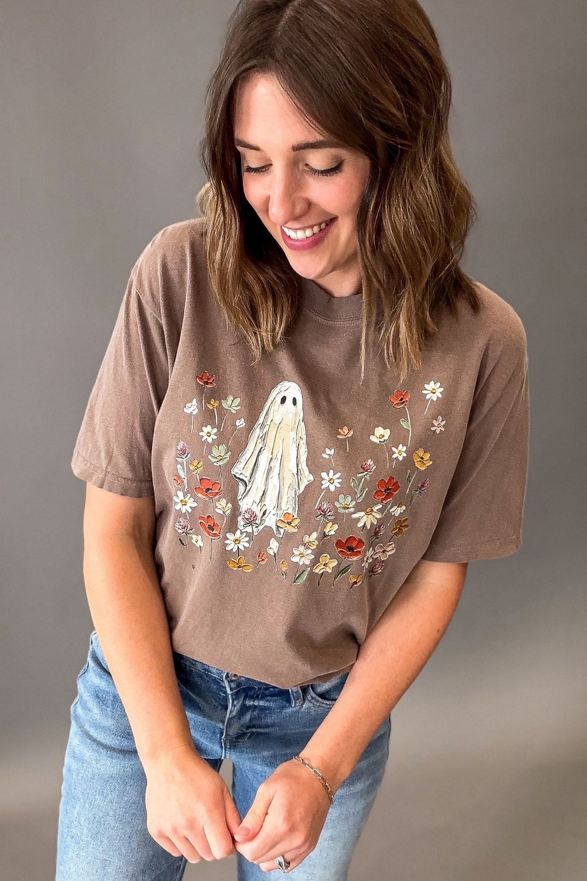 A woman wearing a brown graphic t-shirt with a ghost and floral design, paired with blue jeans, photographed against a neutral gray studio background.