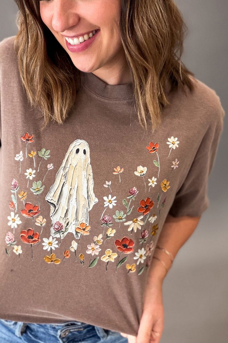 A woman wearing a brown short-sleeve t-shirt with a graphic of a white ghost surrounded by colorful wildflowers, paired with blue jeans.