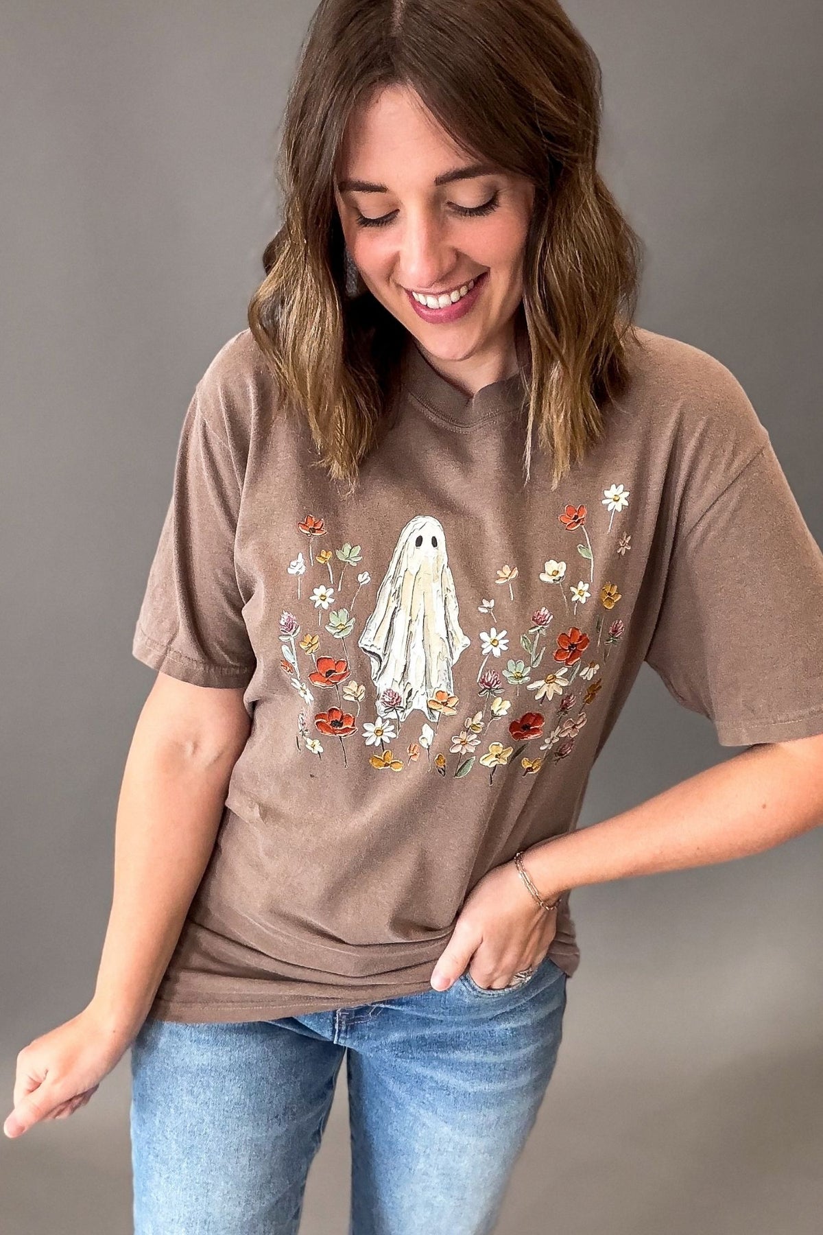 A woman wearing a brown graphic t-shirt with a ghost and wildflowers design, paired with blue jeans, standing against a neutral gray background.