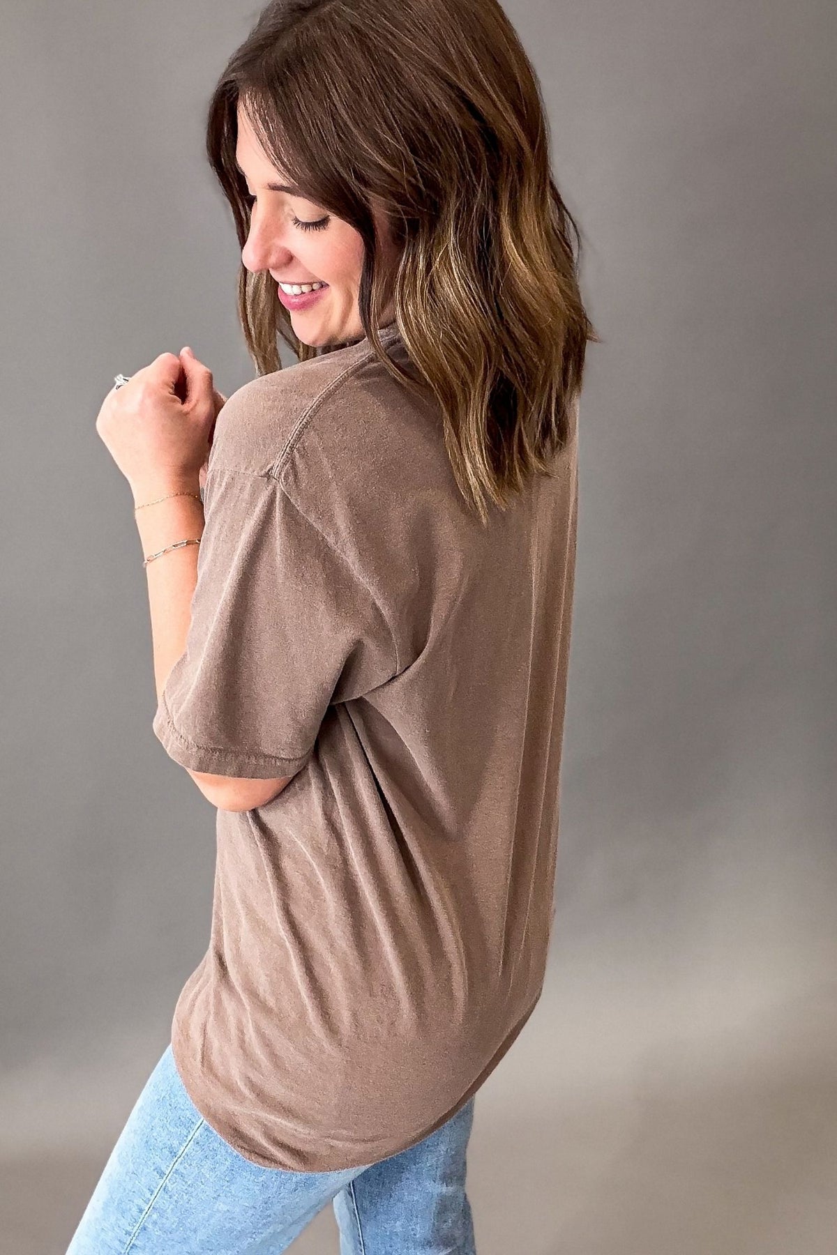 A woman wearing a loose-fitting, short-sleeve espresso-brown t-shirt and light blue jeans, photographed from a side-back angle against a neutral gray studio background.