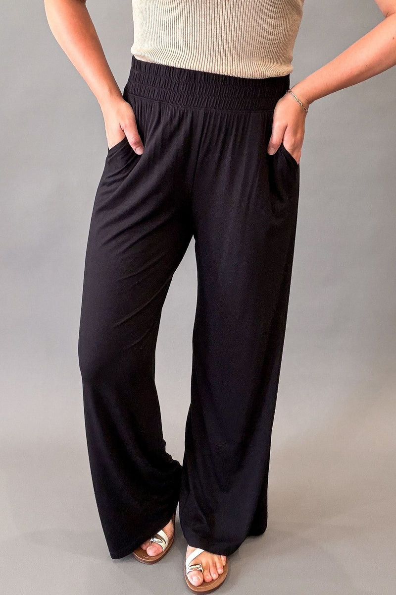 Woman wearing ultra-soft black wide leg pants with a stretchy elastic waistband and front pockets styled with sandals.