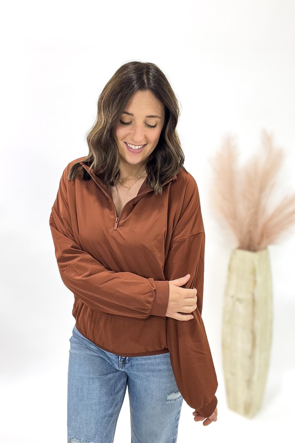 Woman wearing a copper long-sleeve shirt and blue jeans, standing next to a decorative vase with dried plants on a white background.