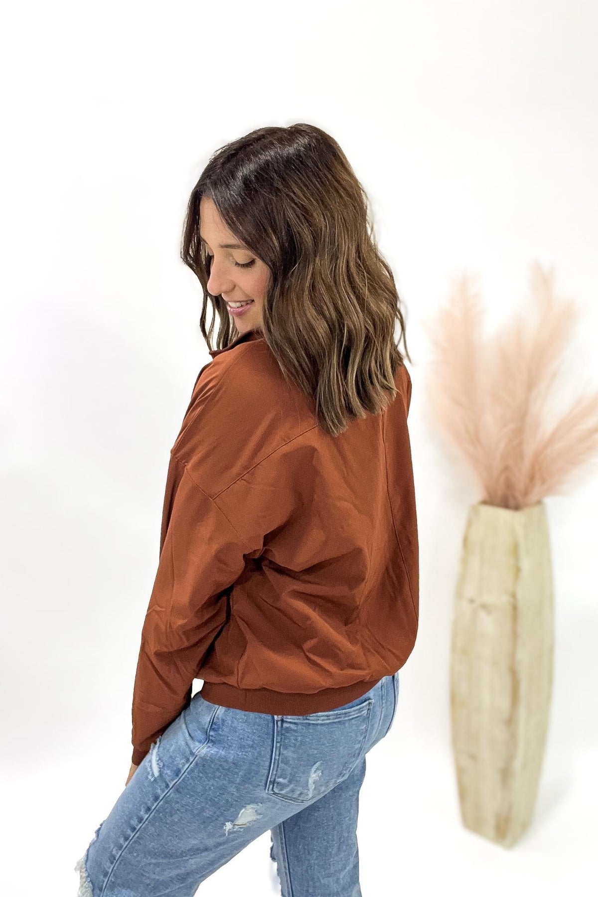 Woman wearing a copper jacket and blue jeans against a white background with a decorative vase.