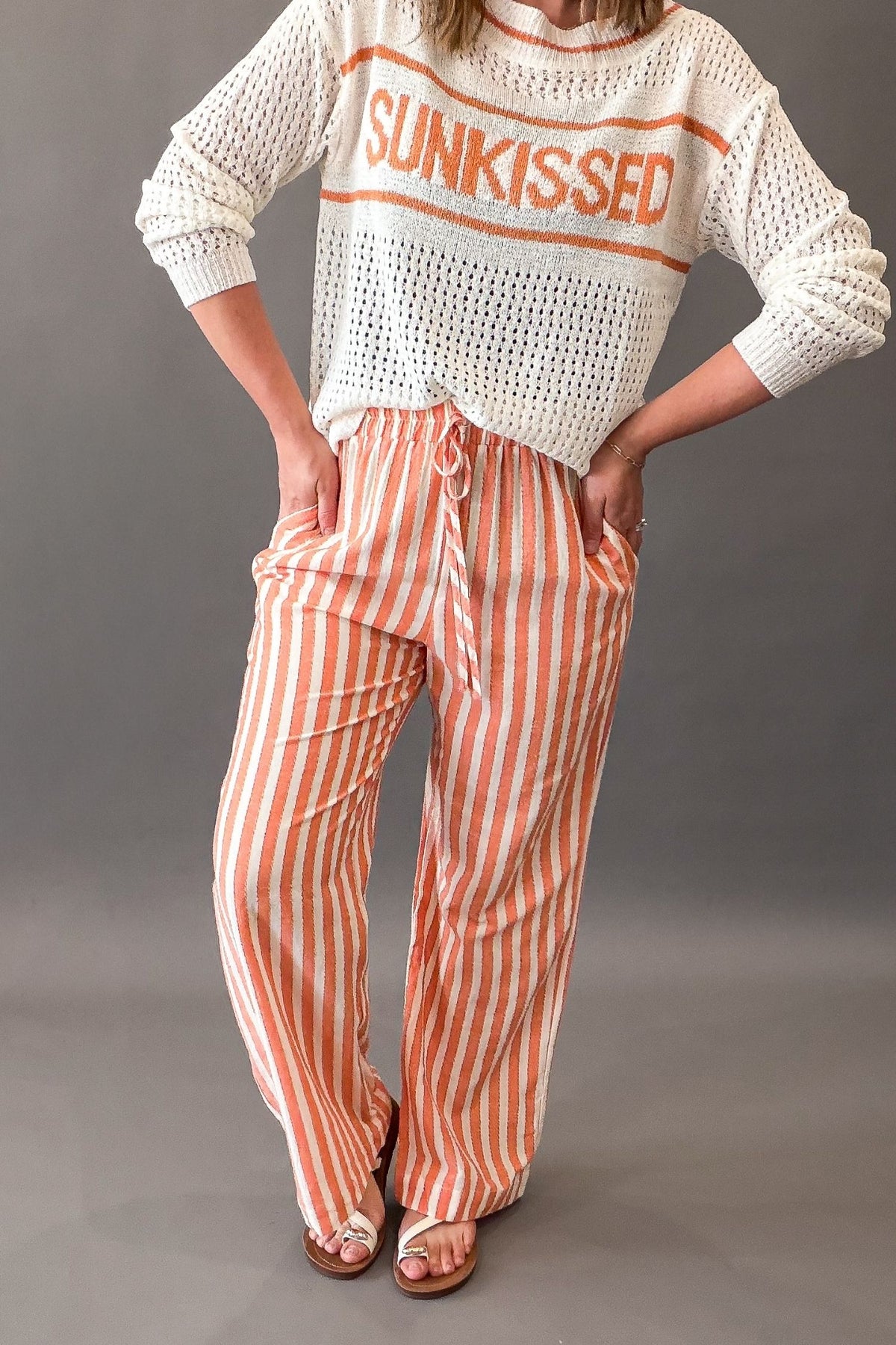 Person wearing a white sweater with 'SUNKISSED' text and coral striped pants on a gray background