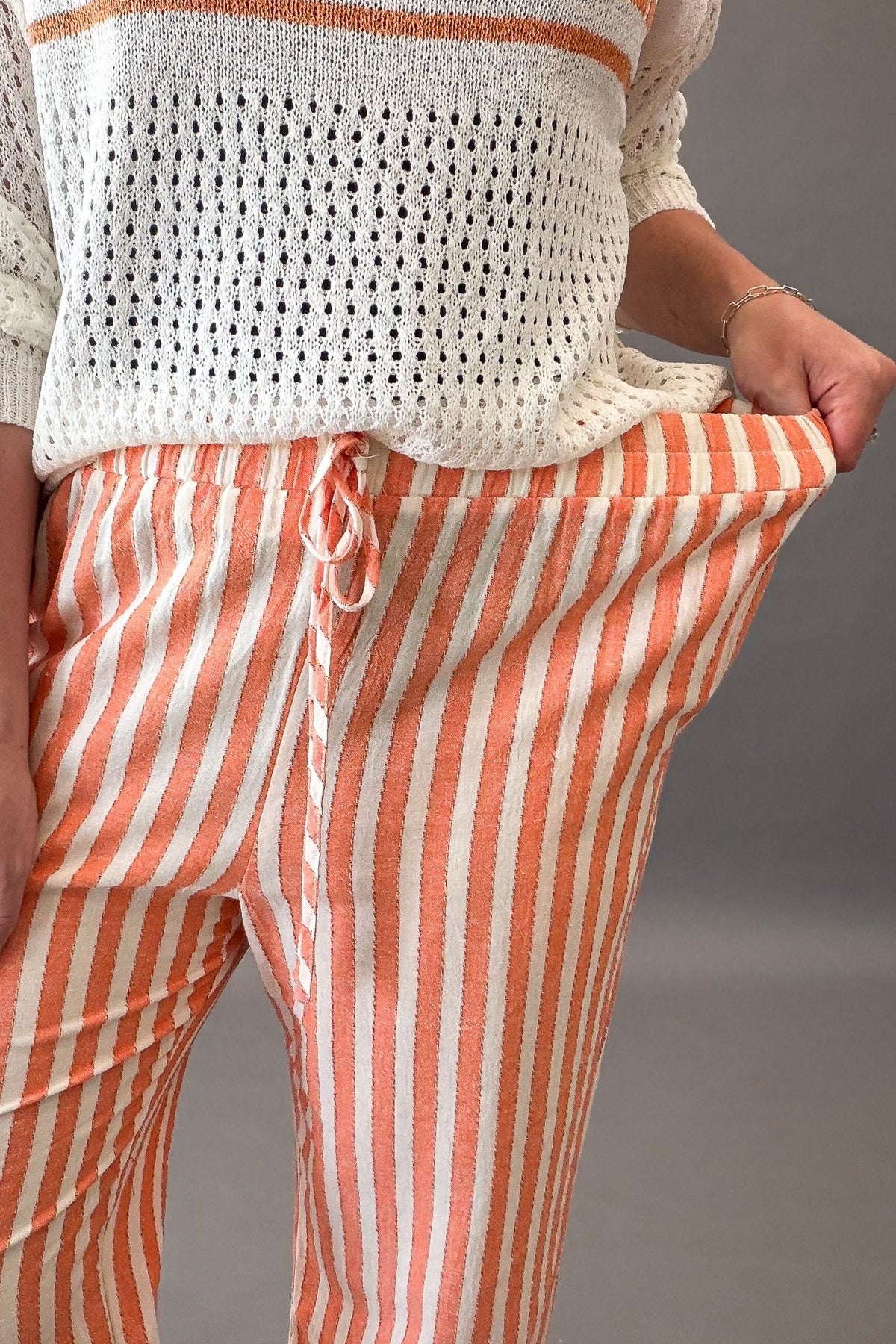 Person wearing a white crochet top and coral and white striped pants on a gray background showing stretch in waistband