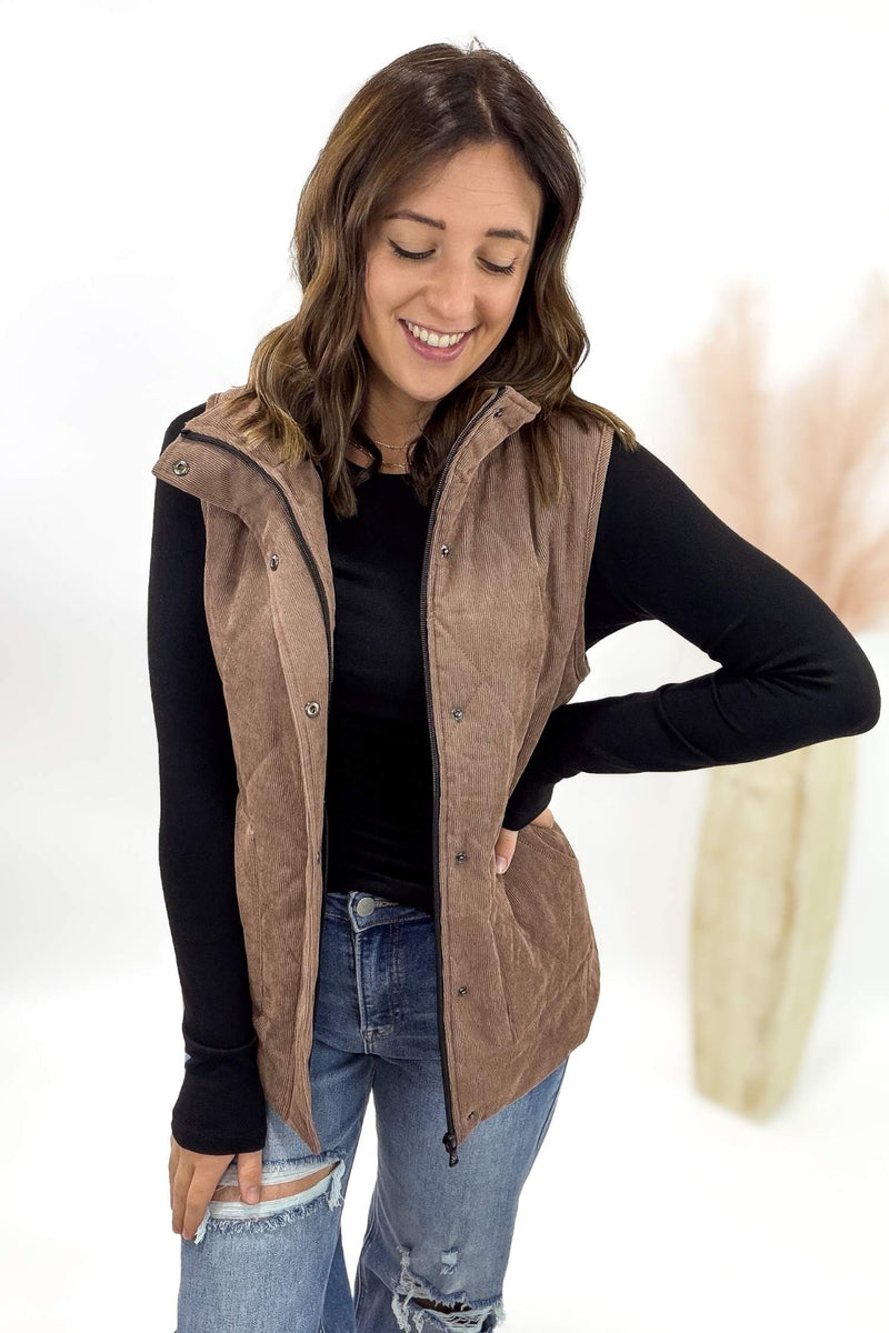 Woman wearing a brown quilted vest over a black long-sleeve shirt and jeans on a white background