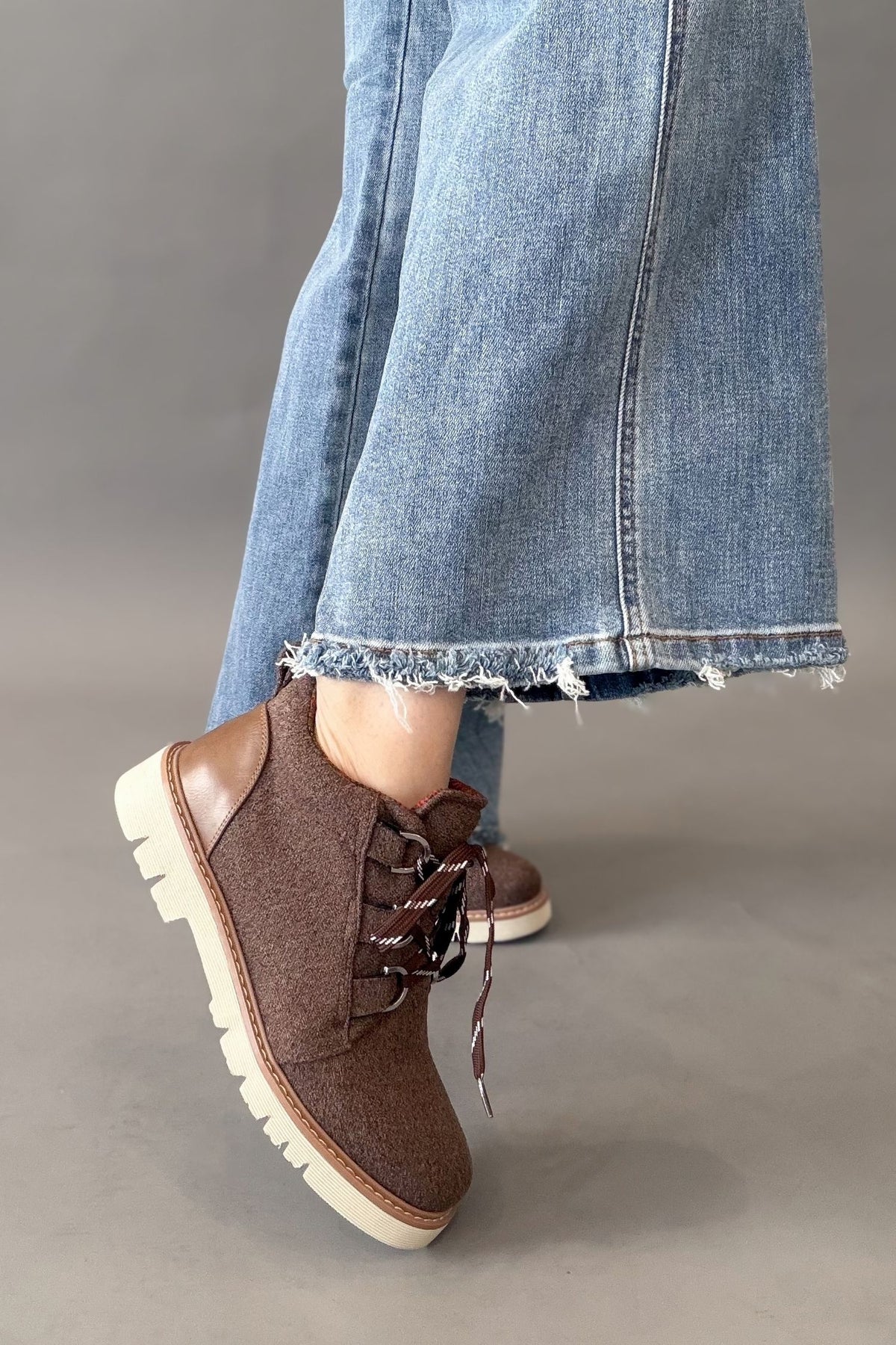 A person wearing brown lace-up ankle boots with thick off-white lug soles, paired with light blue frayed-hem jeans, standing against a neutral gray studio background.