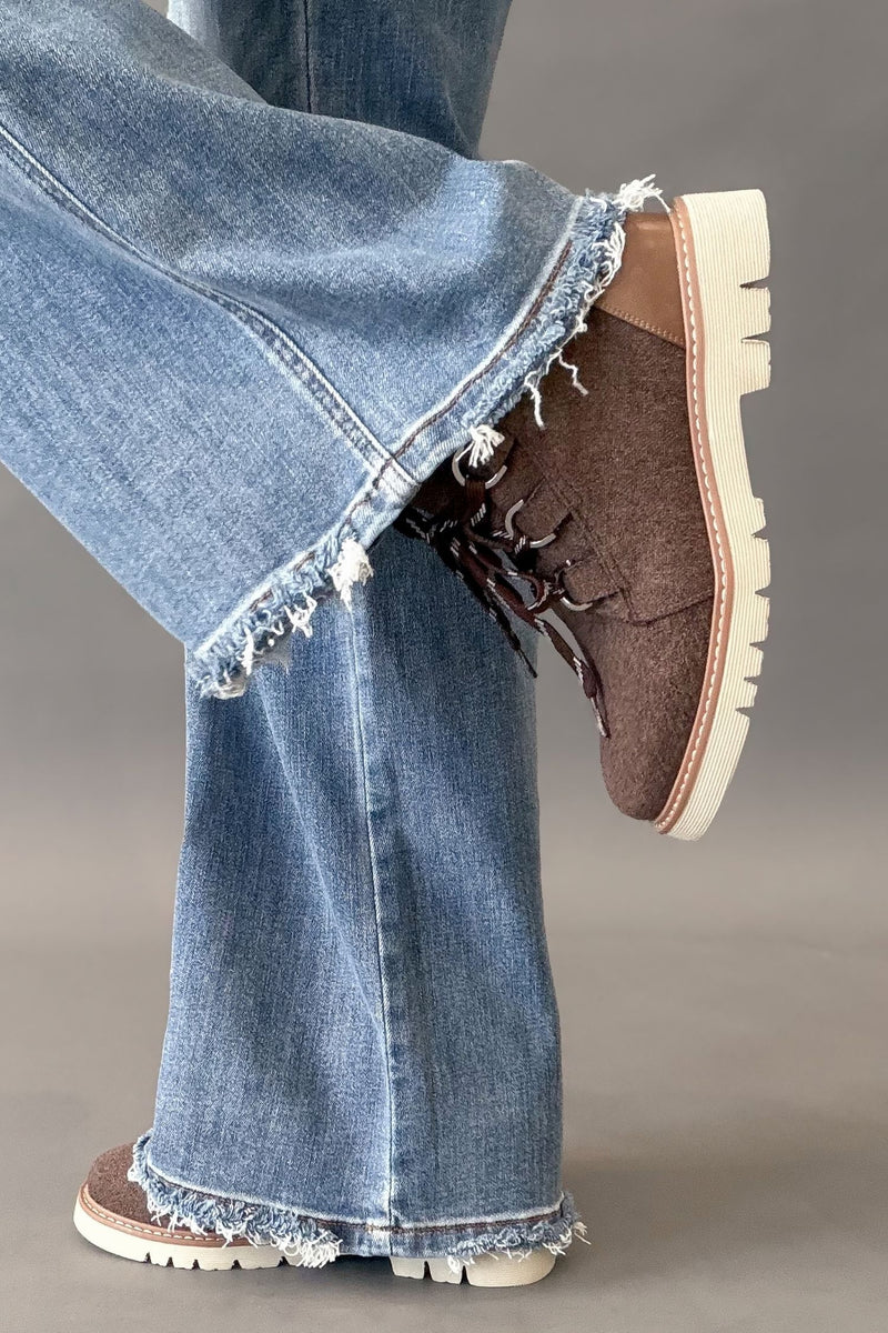 A close-up of a person's legs wearing light blue frayed-hem jeans and brown suede lace-up boots with chunky white lug soles, shown against a neutral gray studio background.