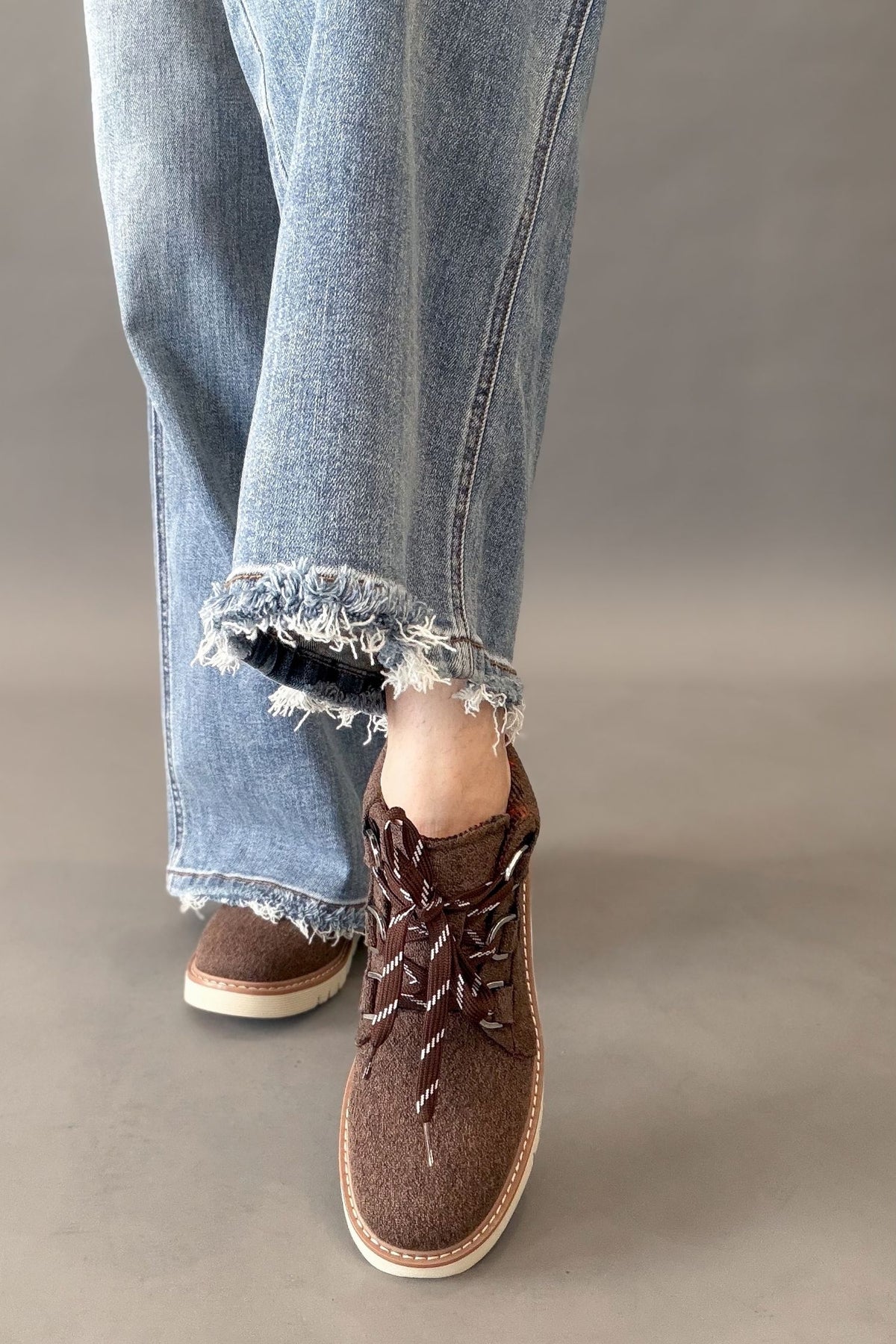 A person wearing light blue frayed-hem jeans and brown textured lace-up shoes with white soles, shown from the knees down against a plain gray studio background.