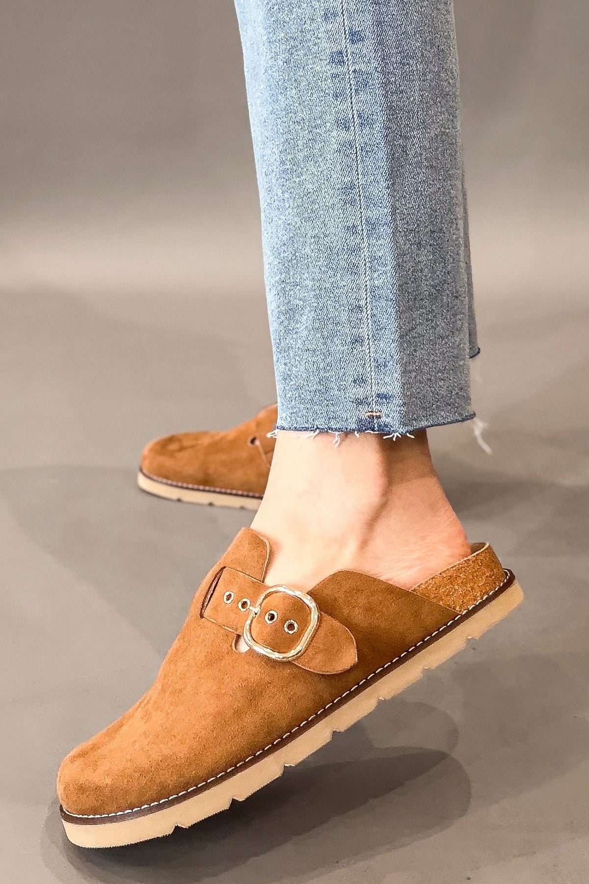 A person wearing light blue jeans and corkys camel faux suede mule clogs with a decorative buckle strap, shown against a neutral gray studio background.
