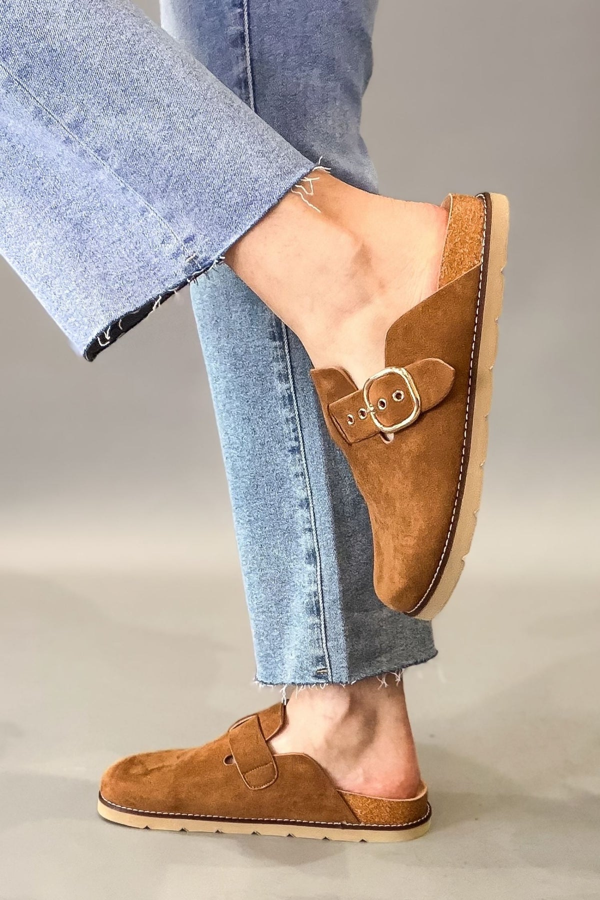 A person wearing light blue jeans and corkys camel suede mule clogs with buckle straps, shown from the knees down against a plain light gray studio background.