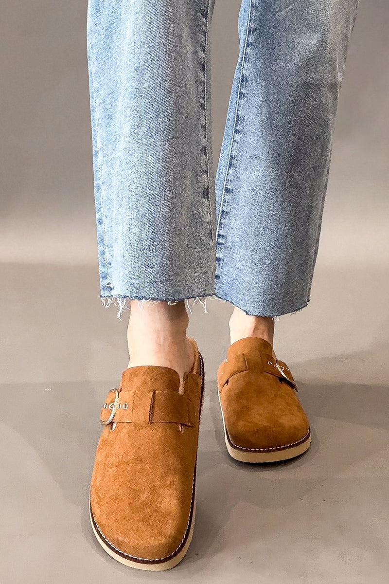 A person wearing light blue frayed-hem jeans and corkys camel faux suede mule loafers with gold-tone buckle details, standing against a neutral gray studio background.