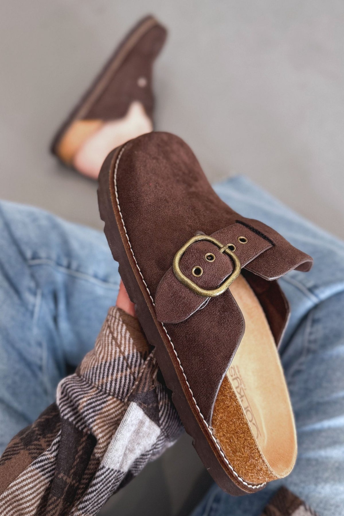 A person wearing brown suede clogs with a gold-tone buckle strap, sitting in blue jeans with a plaid shirt visible.