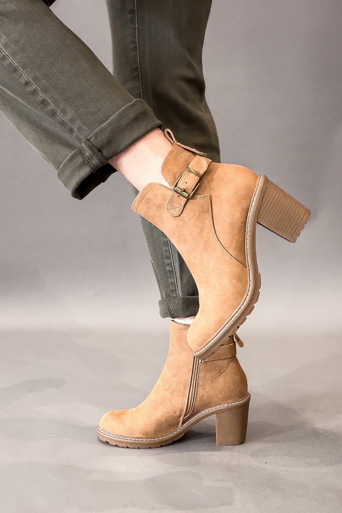 A person wearing camel faux suede ankle boots with a block heel and side buckle strap, paired with olive green cuffed jeans, against a neutral gray studio background.